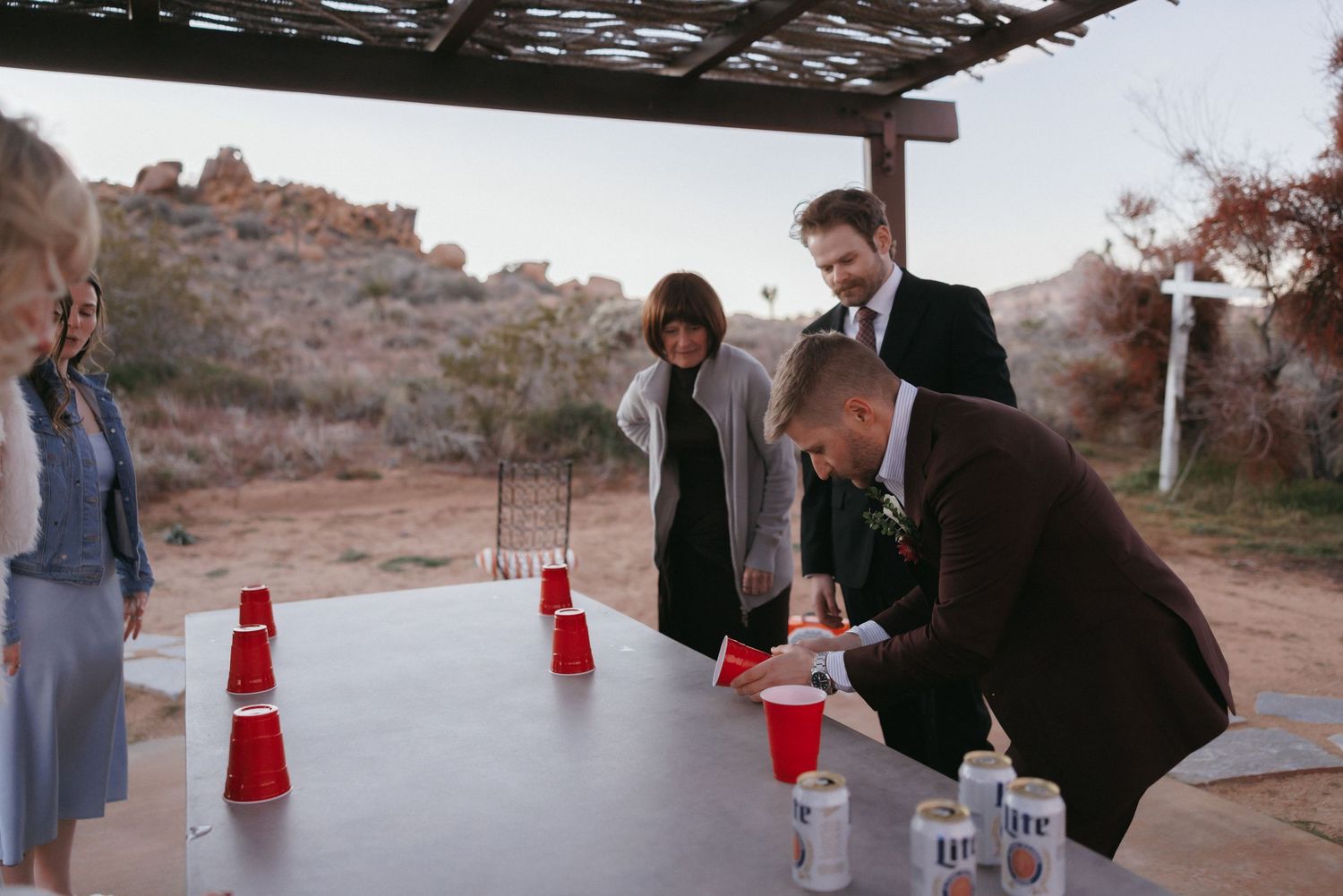 Groom playing beer pong with wedding guests at rustic outdoor desert reception venue.