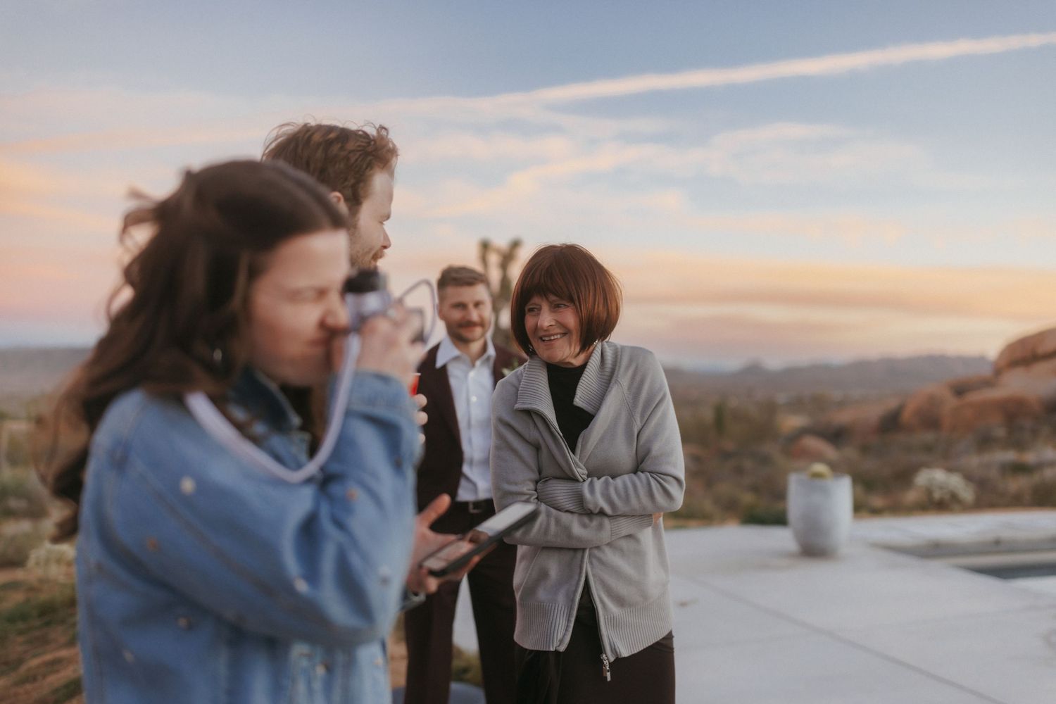 Wedding guests laughing and taking photos at desert venue during colorful sunset celebration.