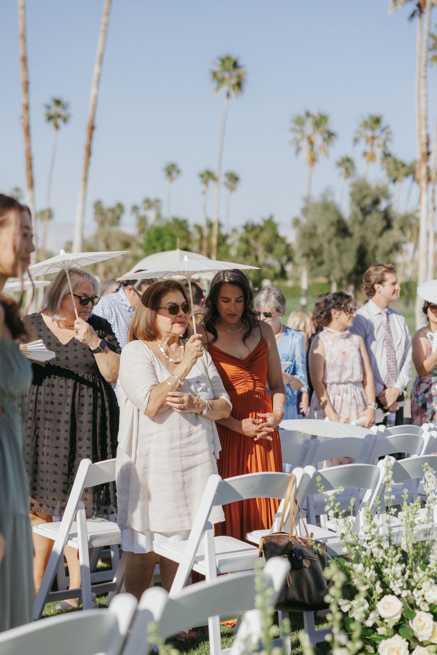 Wedding guests in formal attire arriving at outdoor ceremony with white chairs and palm trees.