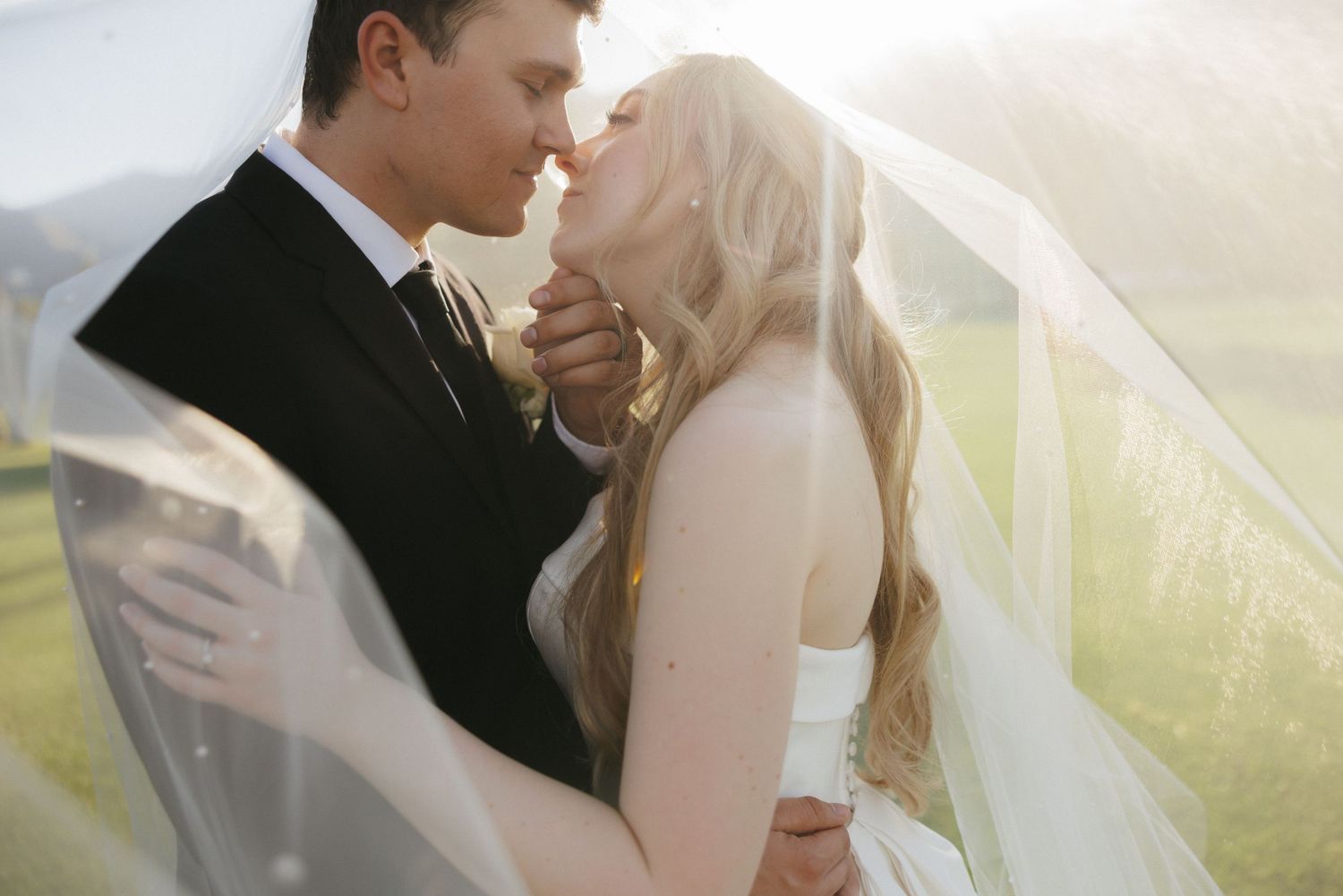 Bride and groom share an intimate moment under a flowing veil with soft golden backlight at outdoor wedding.