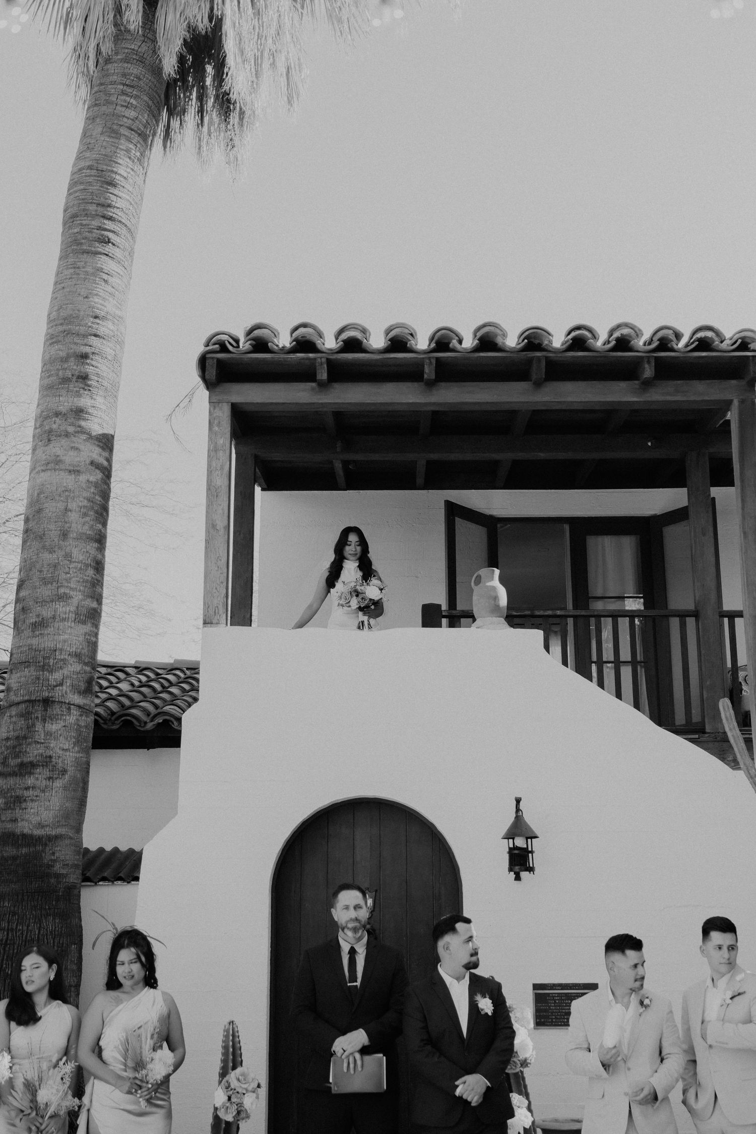 Wedding party members captured in black and white walking outside a Spanish-style building with white stucco walls and palm trees.
