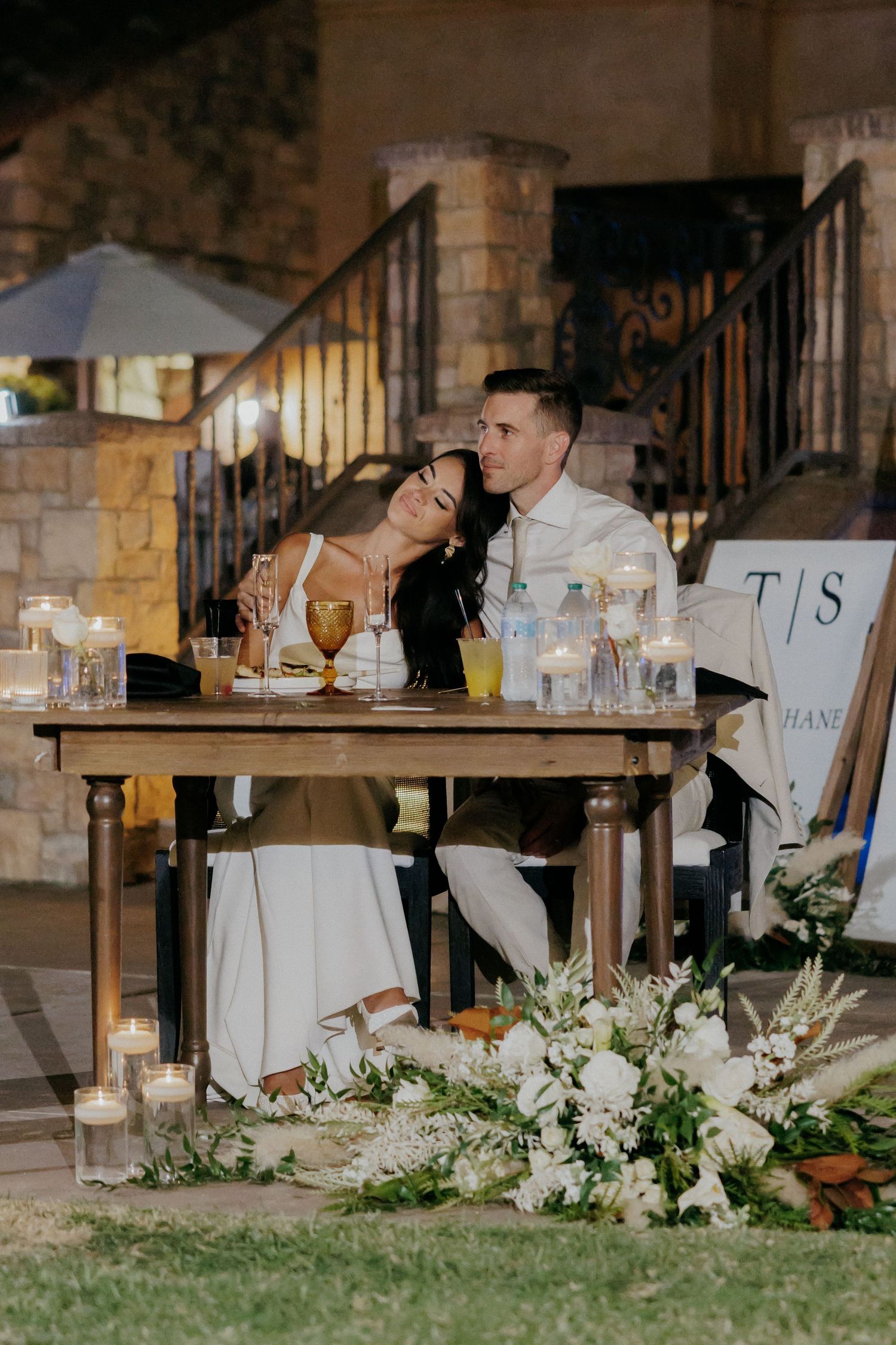Elegant wedding reception table settings with white floral arrangements and glassware in a rustic stone venue.