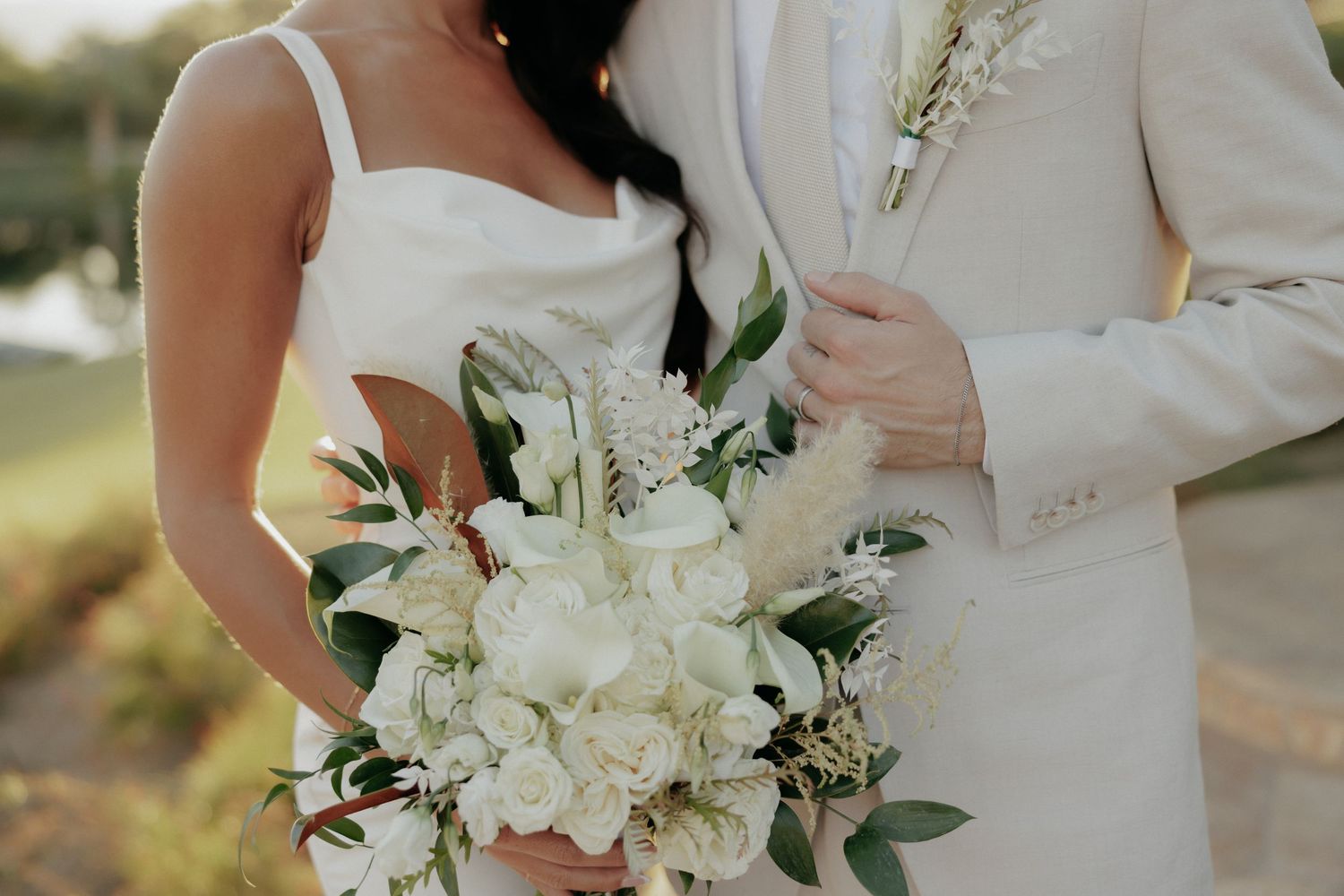 A bride in a white wedding gown holds a romantic bouquet of white roses and greenery in natural outdoor lighting.