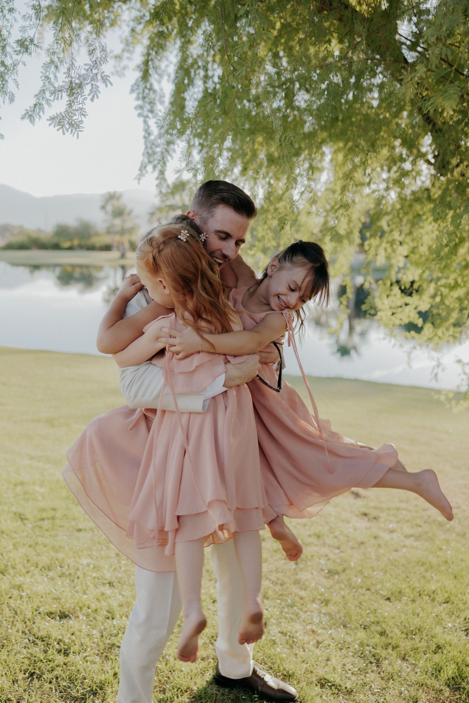 Parent in white embraces and dips a child in pink dress under willow trees by lakeside in playful moment.