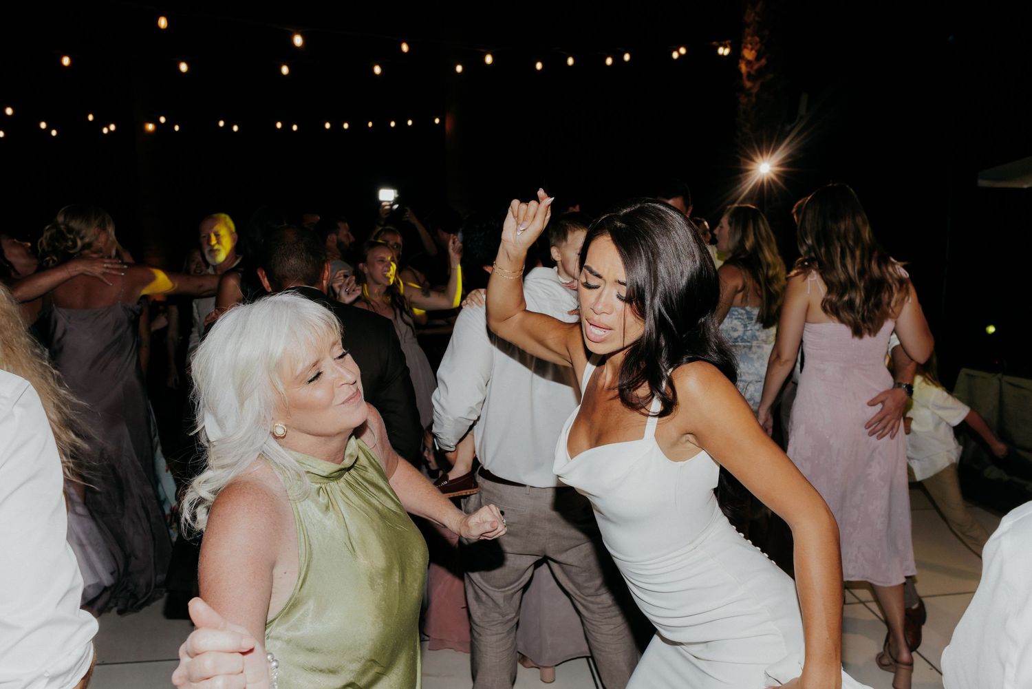 People mingling and dancing at a dimly lit evening event with twinkling lights above.