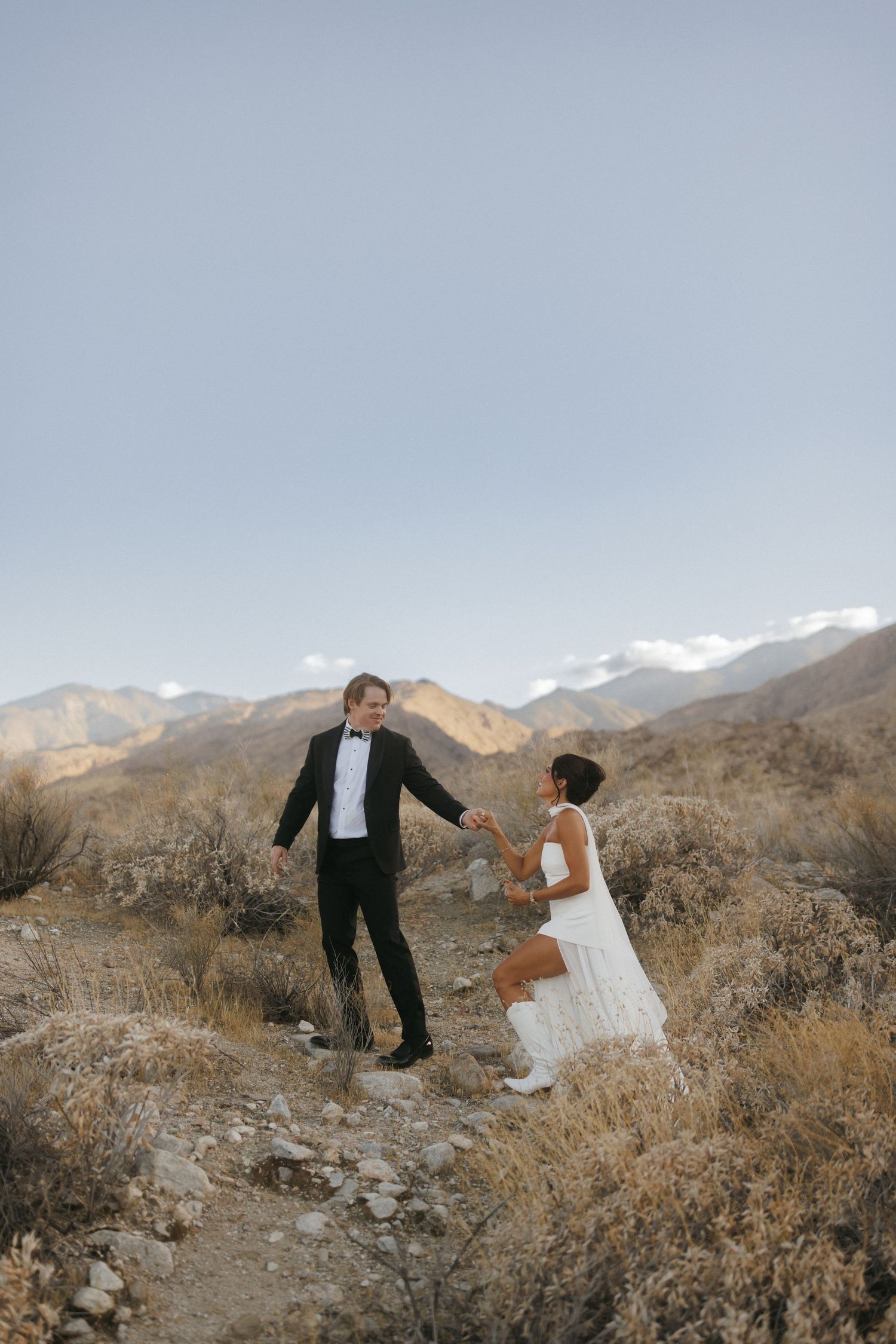 Couple in wedding attire holding hands on a desert trail with mountain backdrop.