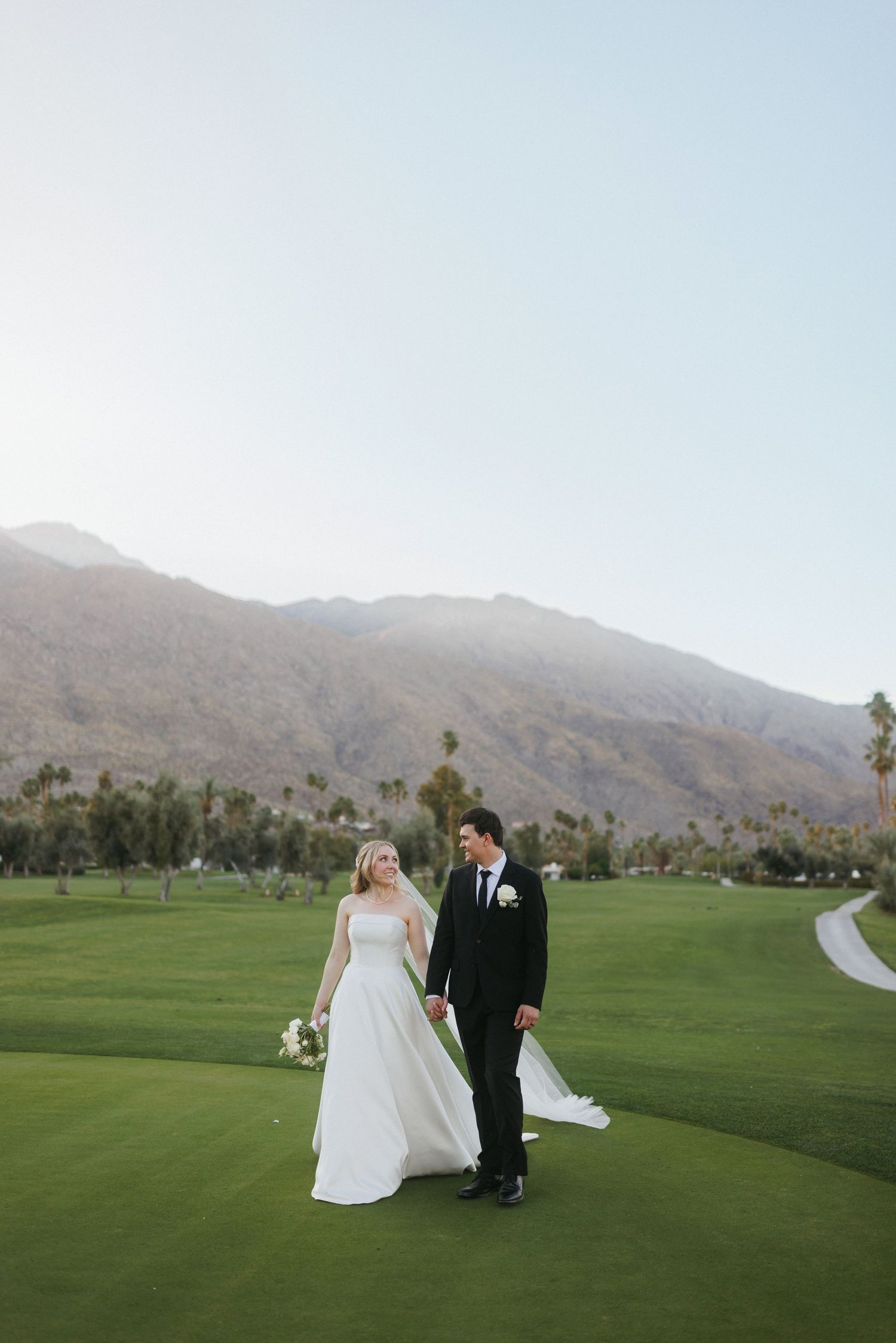 Bride and groom walk hand in hand on lush green golf course with mountain views.