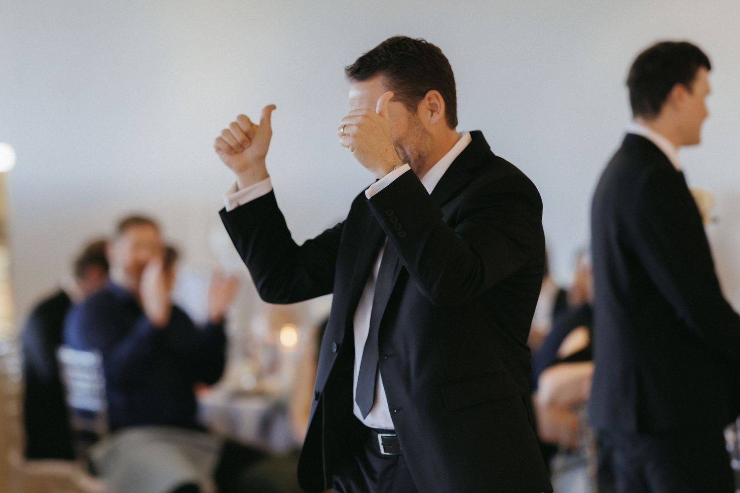 Guest in black suit celebrates with thumbs up during lively wedding reception.
