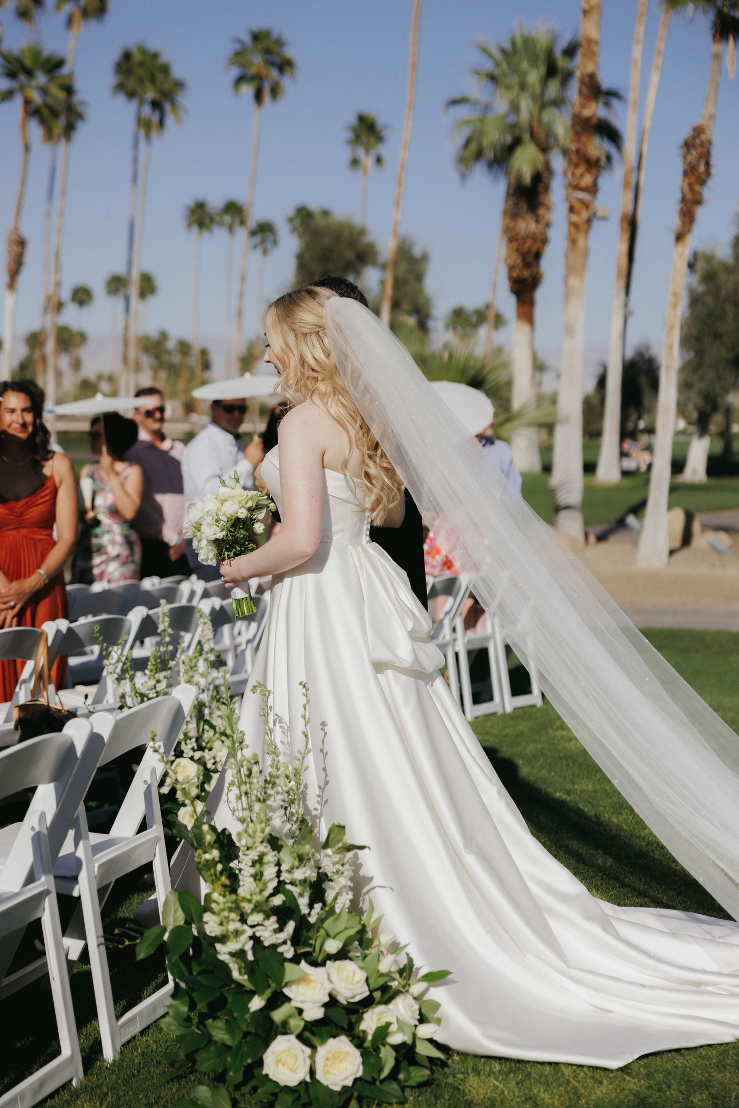 Bride in white gown and veil walks down outdoor aisle lined with white flowers and palm trees.
