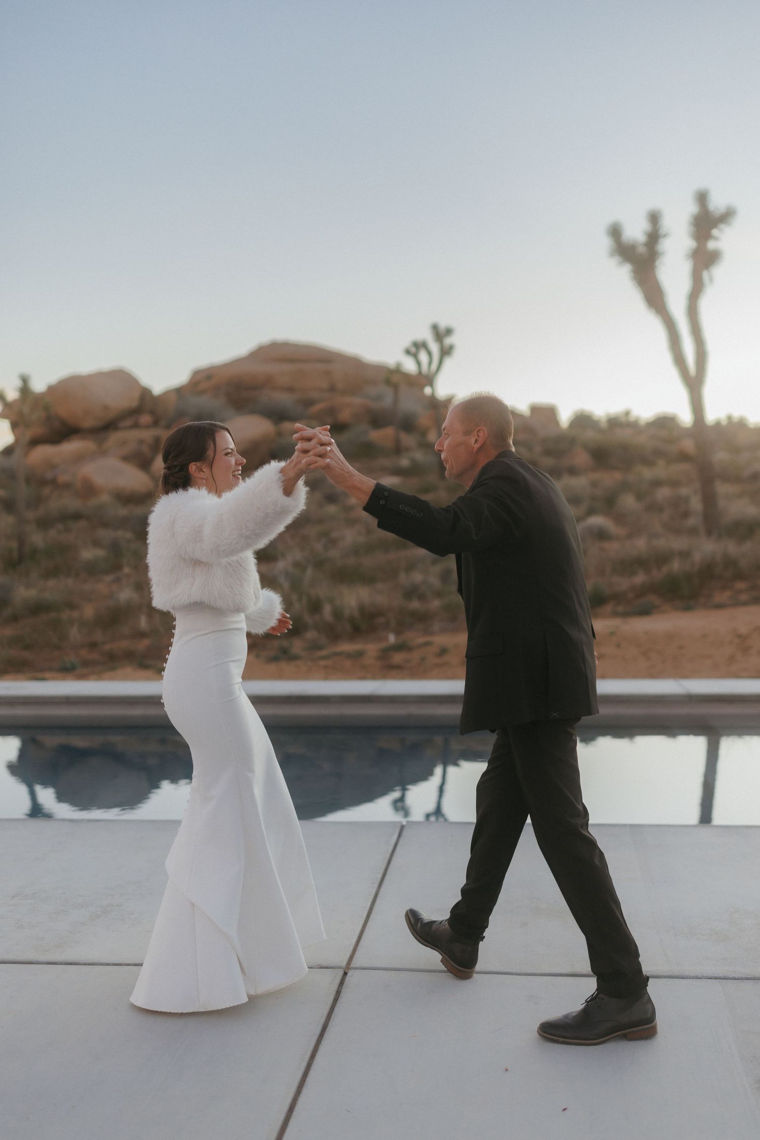 Bride and older man dancing by pool at dusk with Joshua trees and boulders behind.