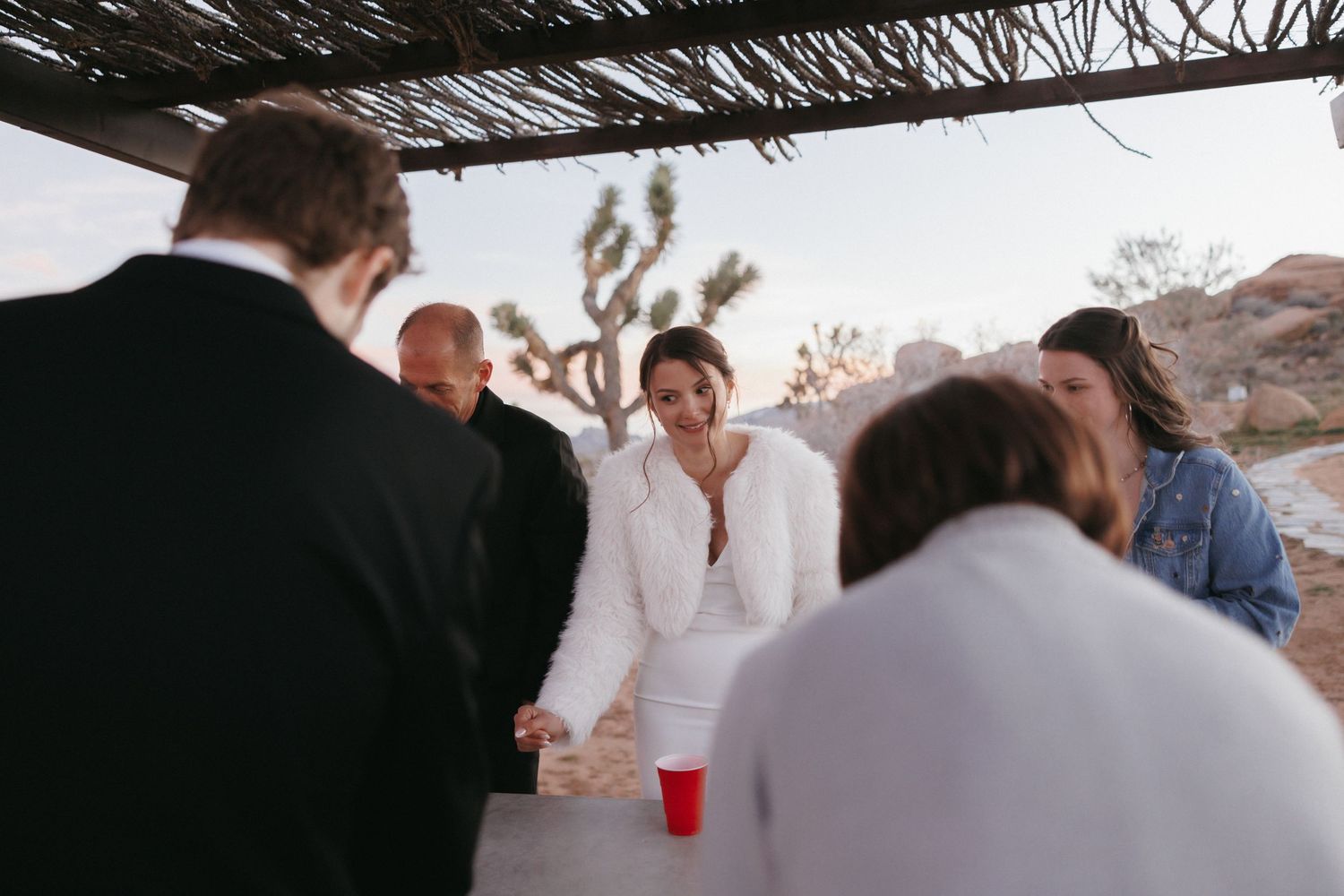 Bride in white fur wrap laughs with wedding guests gathered under a pergola at Joshua Tree reception.