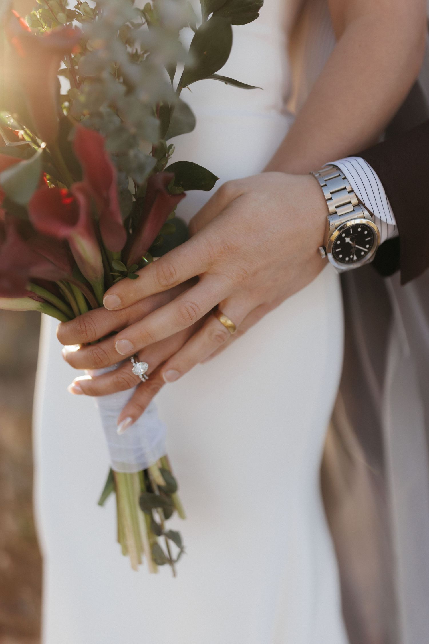 Close-up of bride and groom's hands holding red calla lily bouquet showing engagement ring and watch.