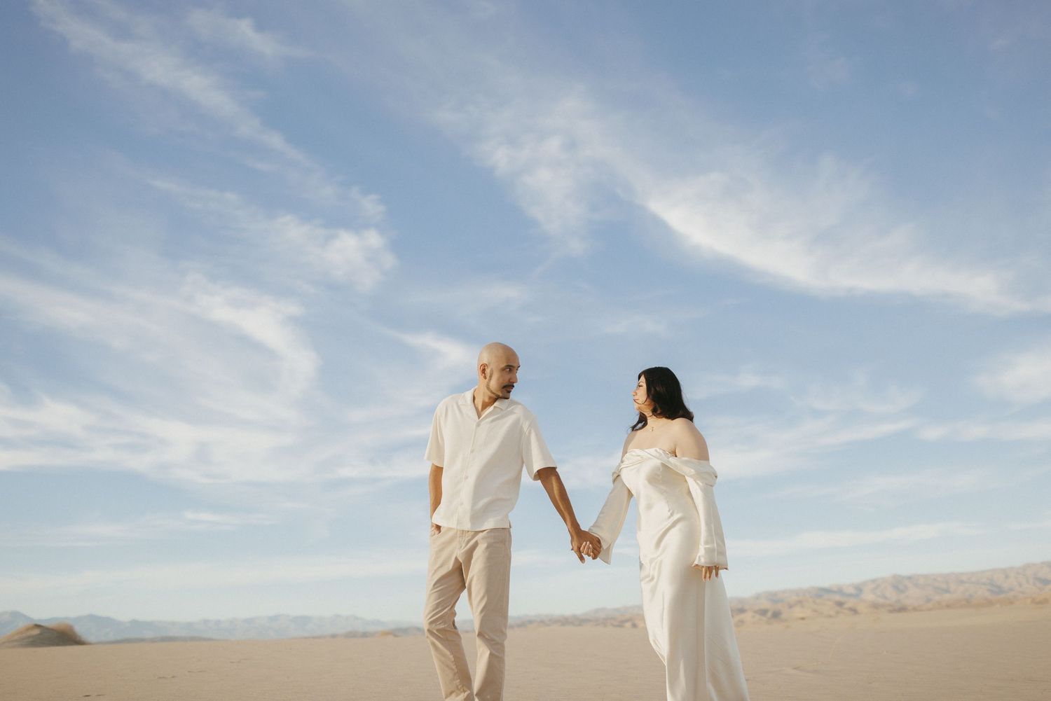 Couple in white outfits holding hands on sandy desert dunes under a blue sky with wispy clouds.