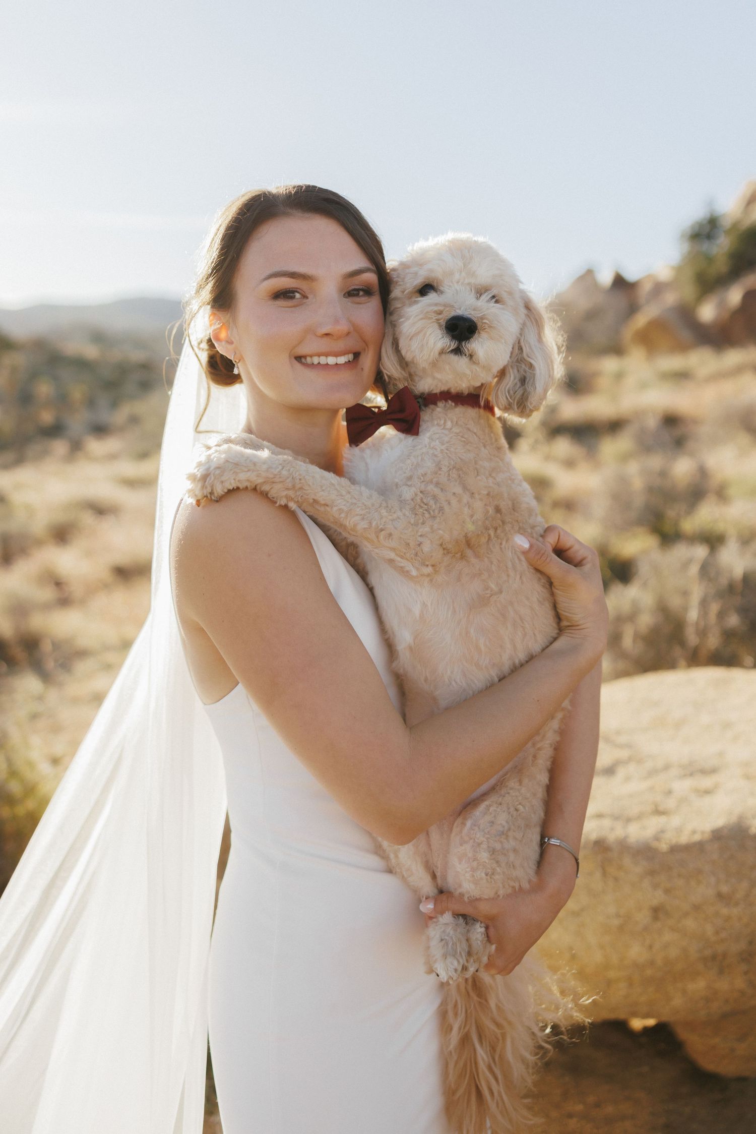 Smiling bride in white gown and veil holds fluffy dog wearing red bow tie in desert landscape.