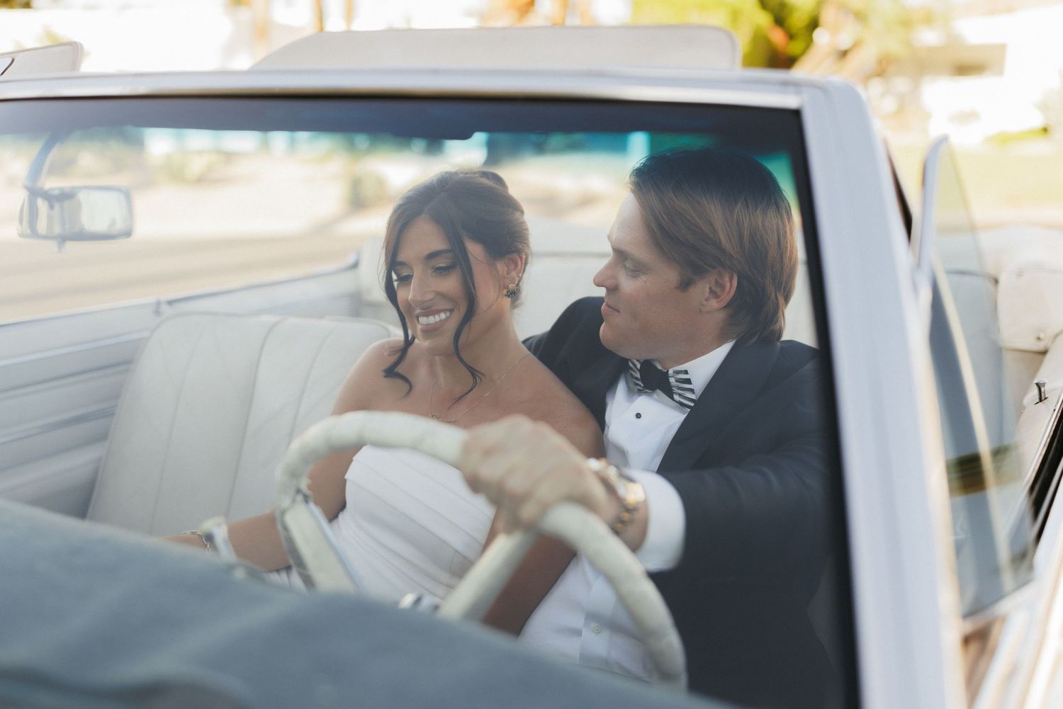 Bride and groom smiling together in a classic white convertible car on their wedding day.