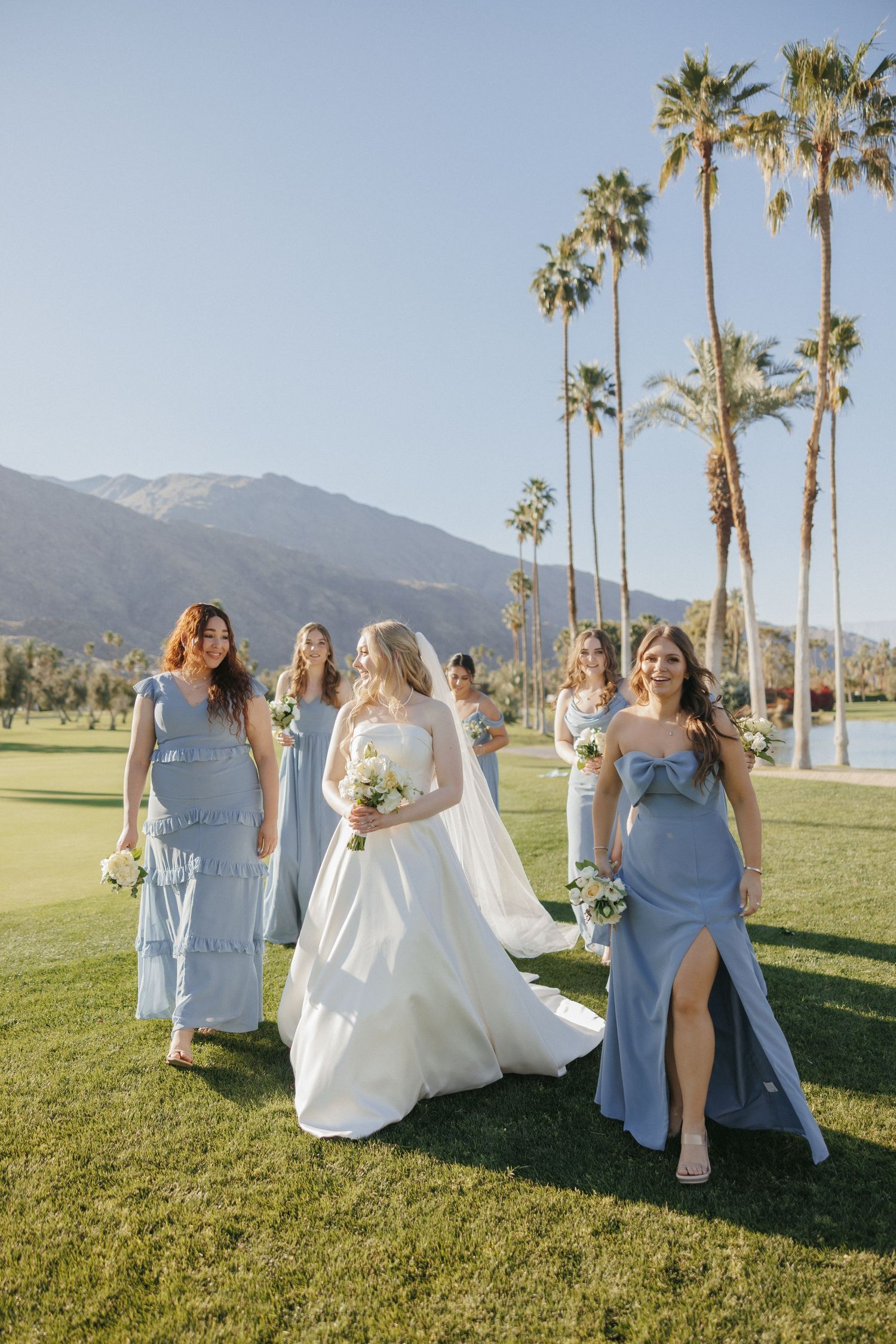 Bride in white gown walking with bridesmaids in blue dresses on golf course with palm trees and mountains.