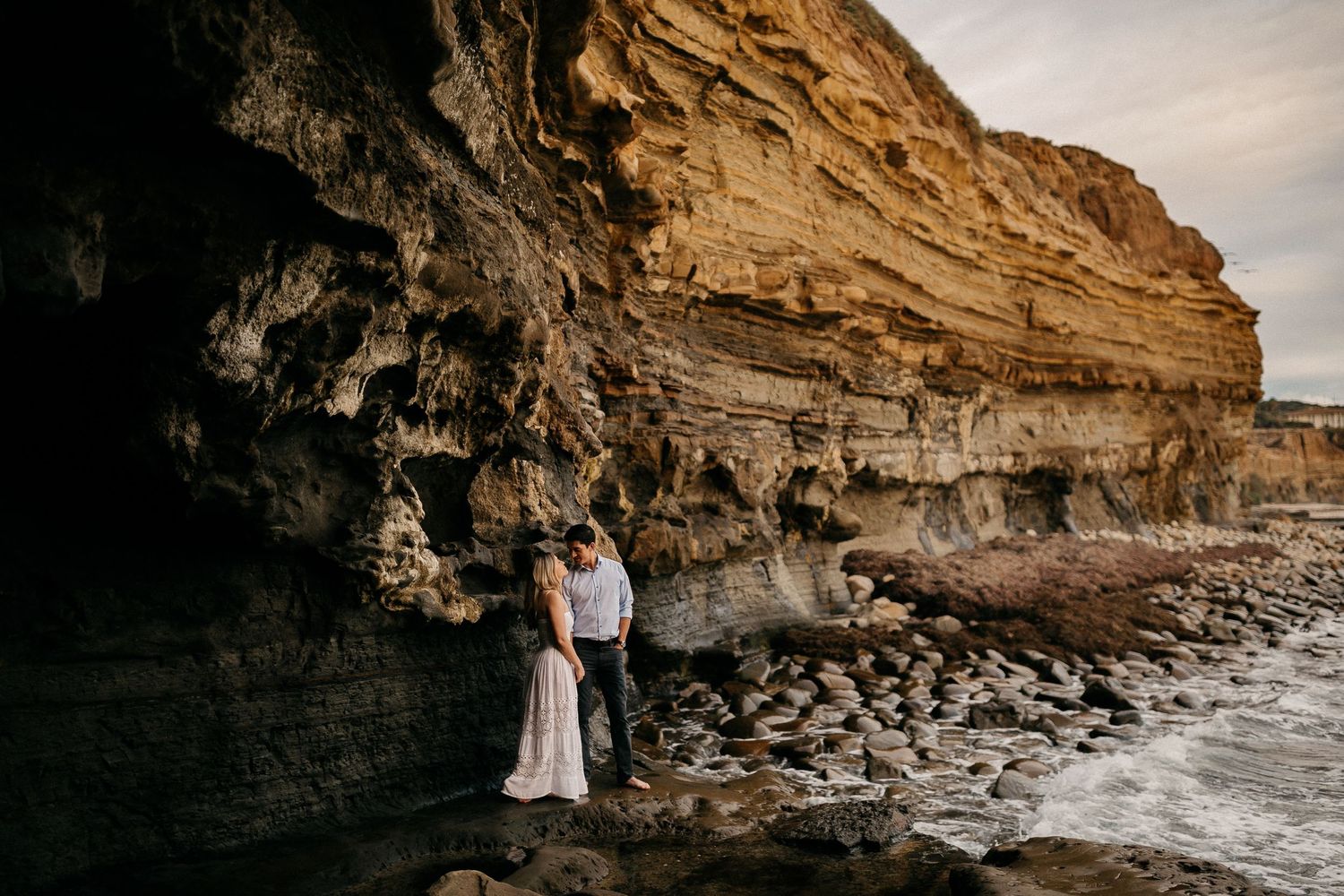 Couple embracing near rocky coastal cliffs at sunset as waves crash against the shoreline during golden hour.
