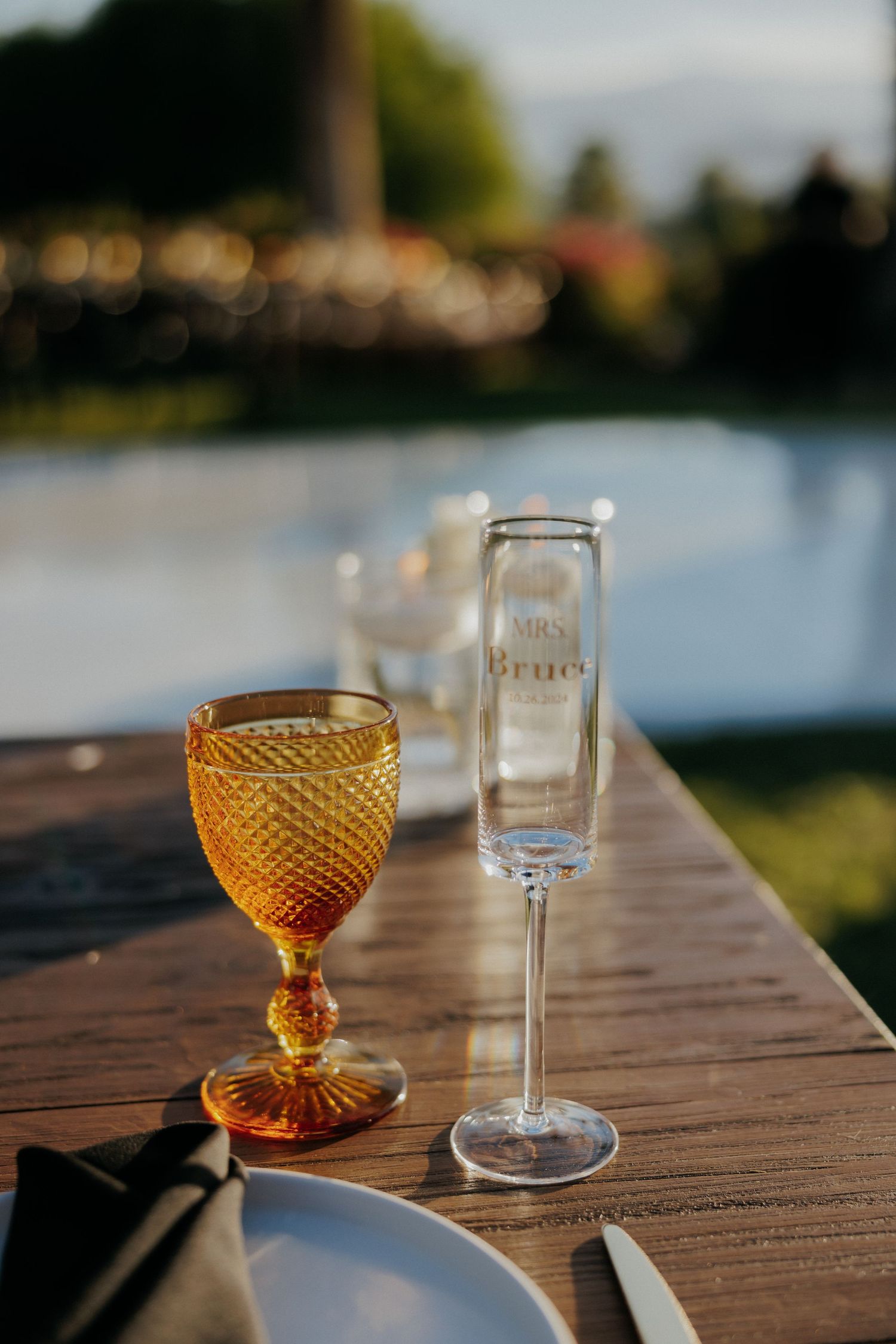 Amber wine glasses and clear shot glasses arranged on a rustic wooden table overlooking a serene lake at sunset.