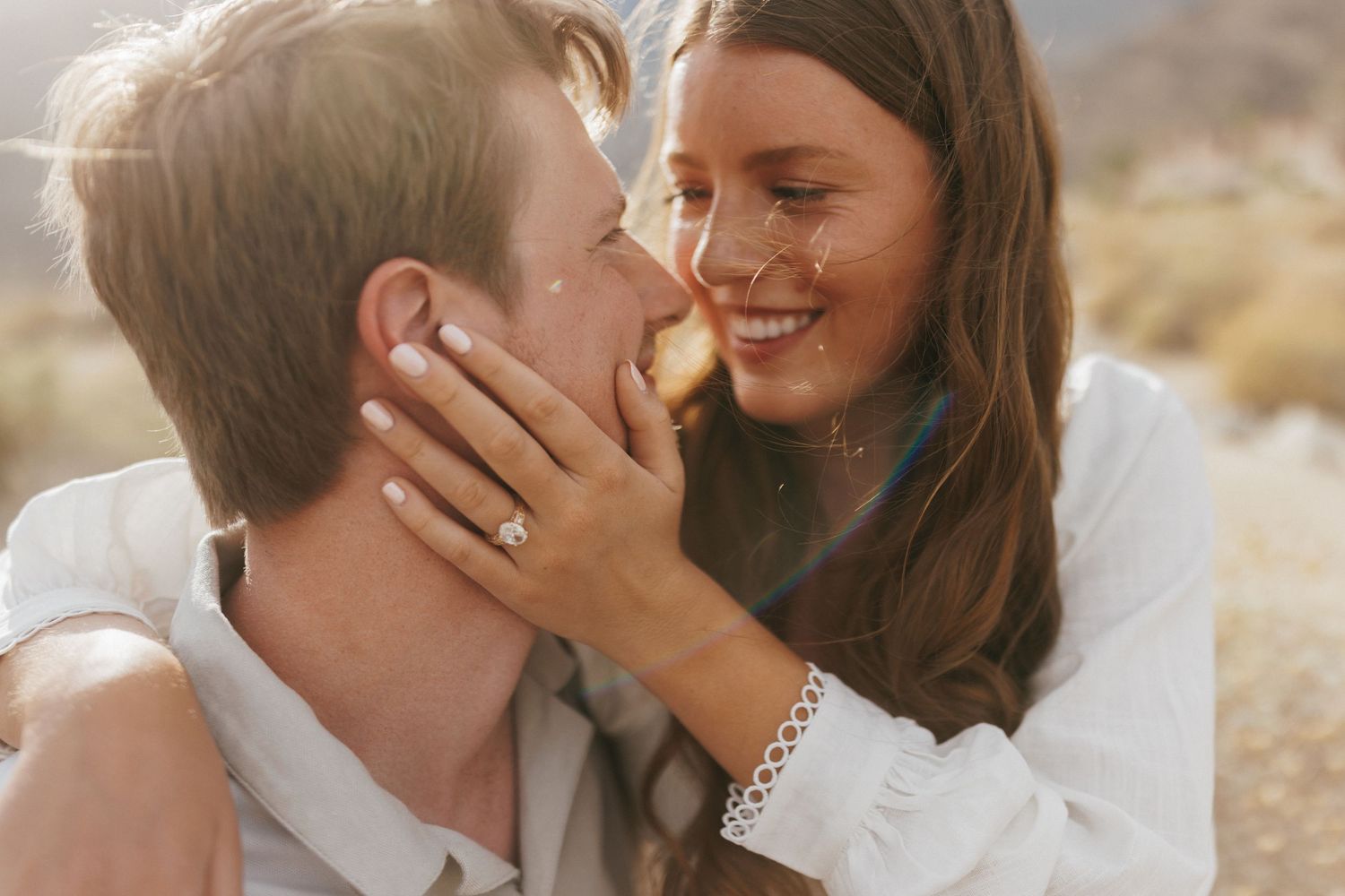 Couple sharing a romantic moment outdoors, woman smiling with engagement ring visible, sun-kissed desert backdrop.