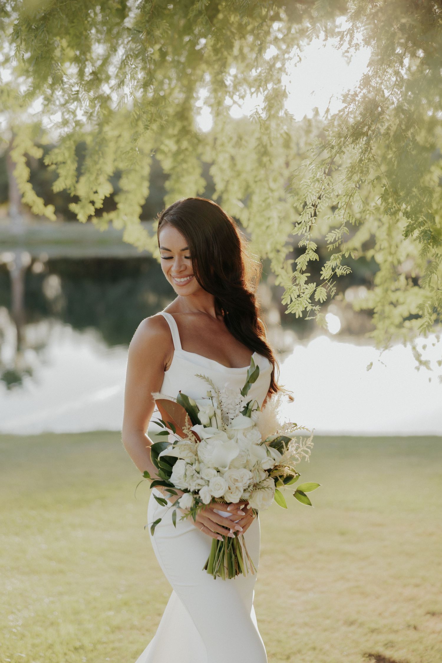 A bride in a fitted white dress holds an elegant bouquet of white flowers under sunlit willow trees.