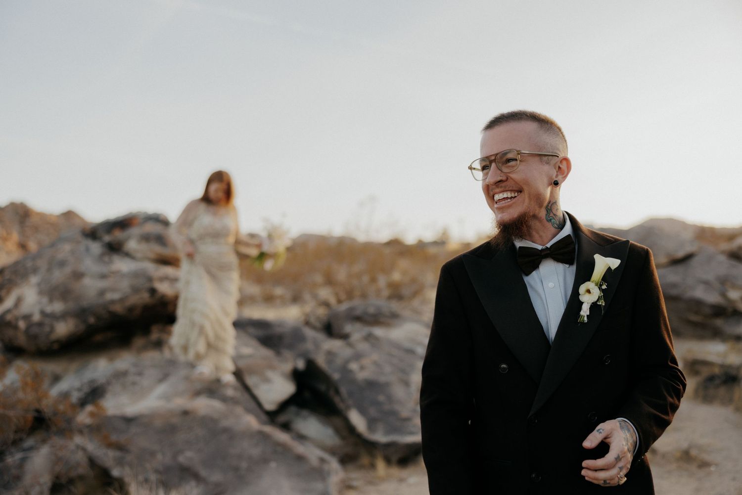 A wedding scene with someone in a black tuxedo and bowtie standing on rocky terrain at dusk.