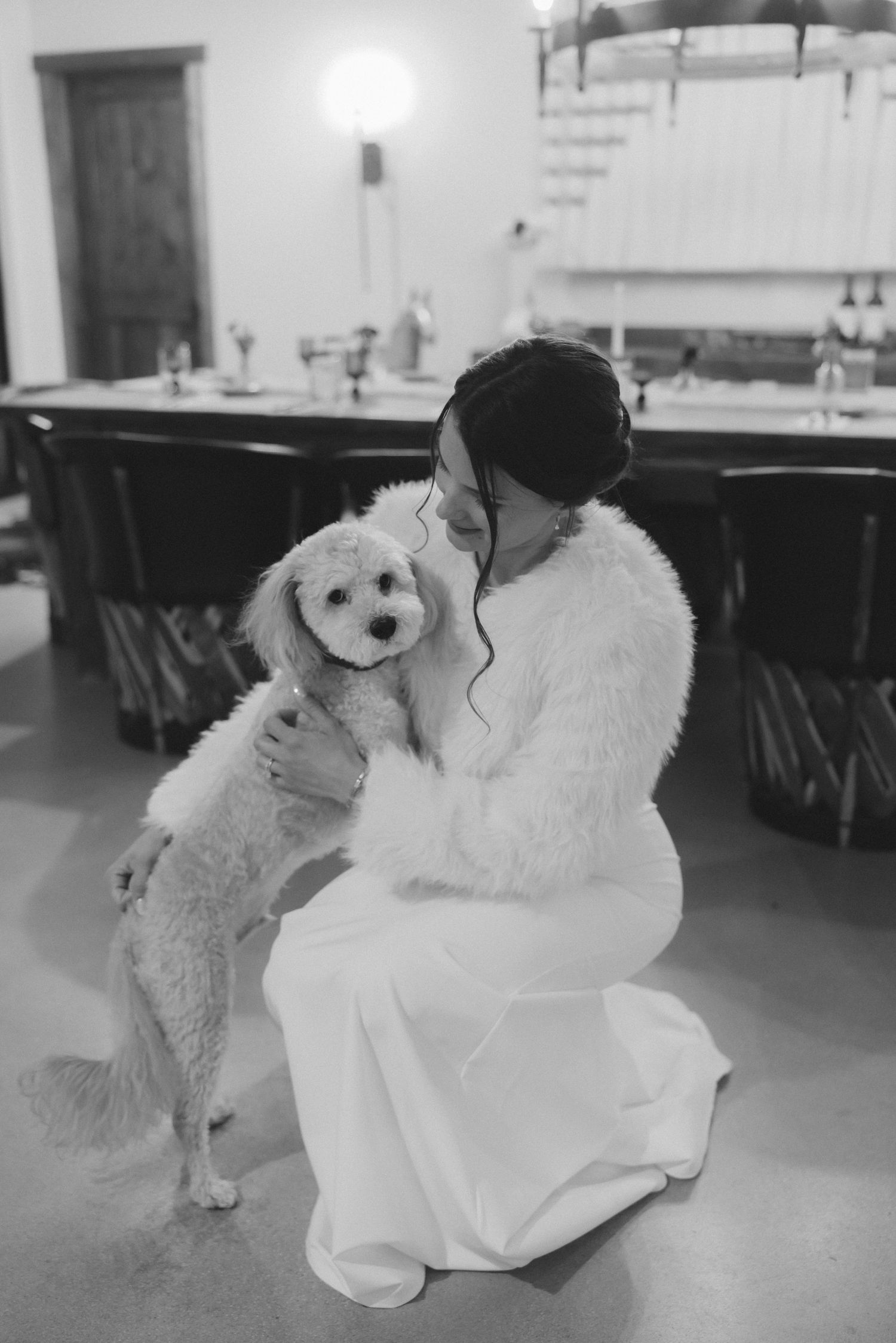 Bride in white gown cuddles fluffy white poodle in black and white photo.
