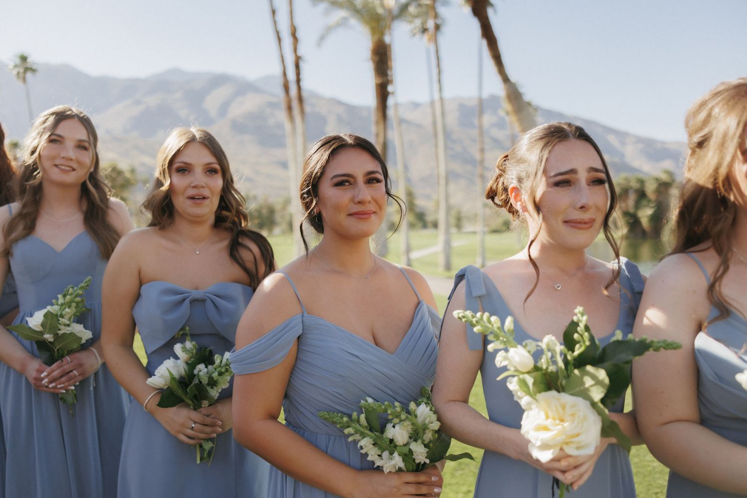 Bridesmaids in matching dusty blue off-shoulder gowns hold white bouquets with palm trees and mountains behind them.