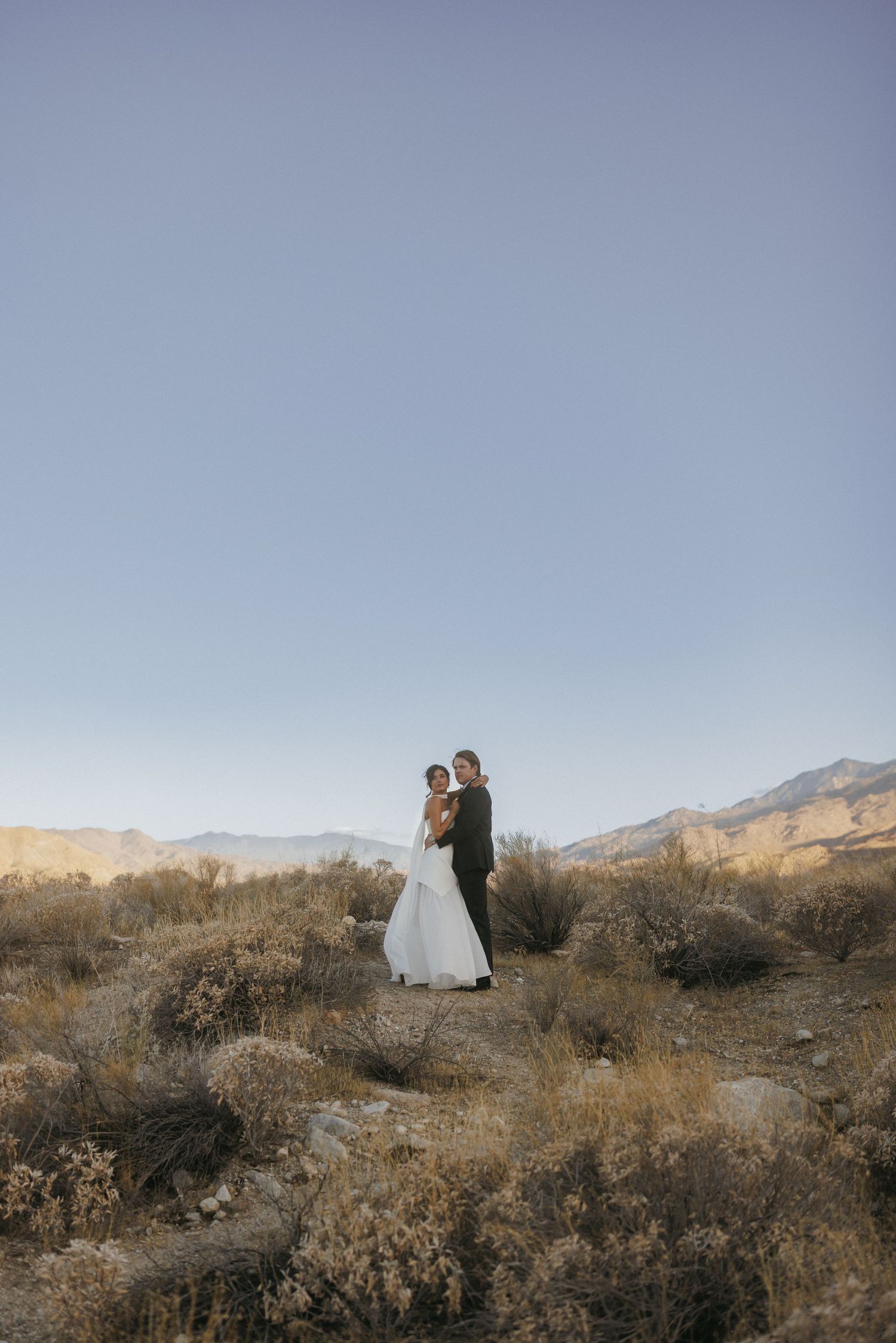 Bride and groom embracing on a desert hilltop with mountain backdrop and clear blue sky at golden hour.