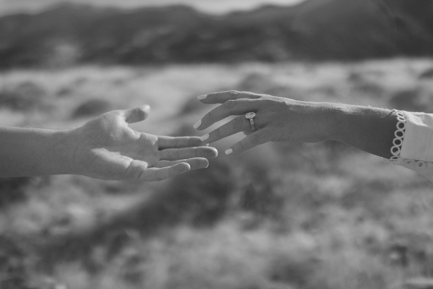 Black and white close-up of two hands reaching toward each other, one wearing an engagement ring.