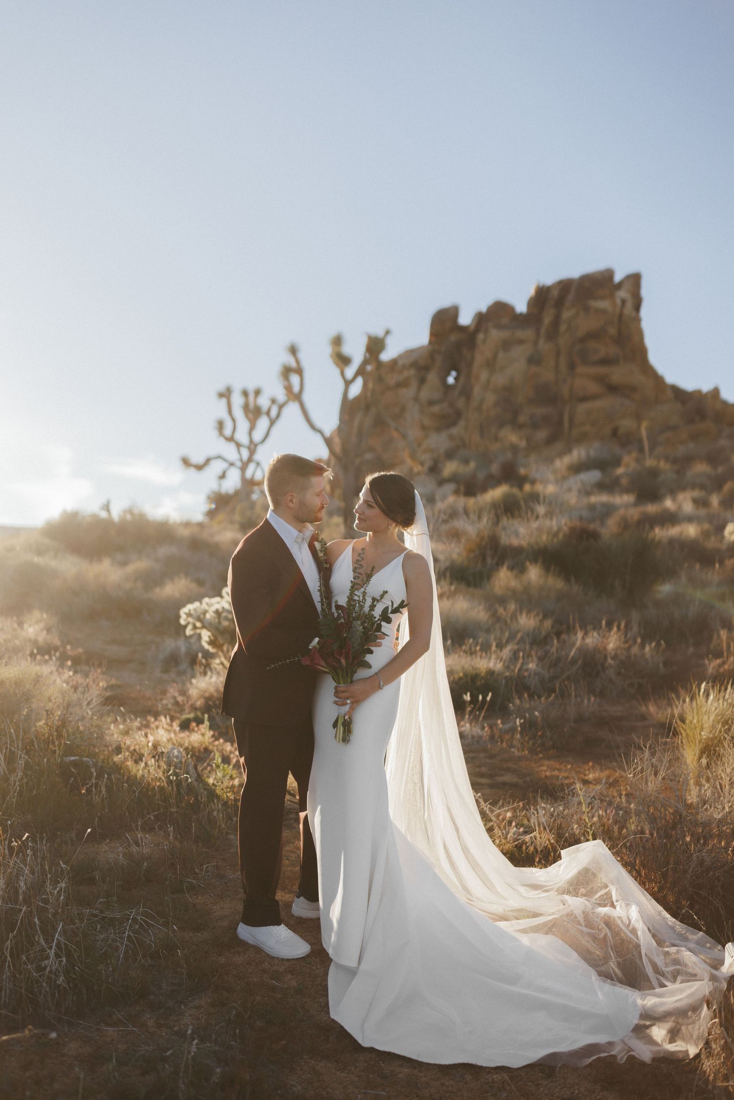 Bride and groom share a romantic moment at golden hour in Joshua Tree with glowing sunlight and rock formations.