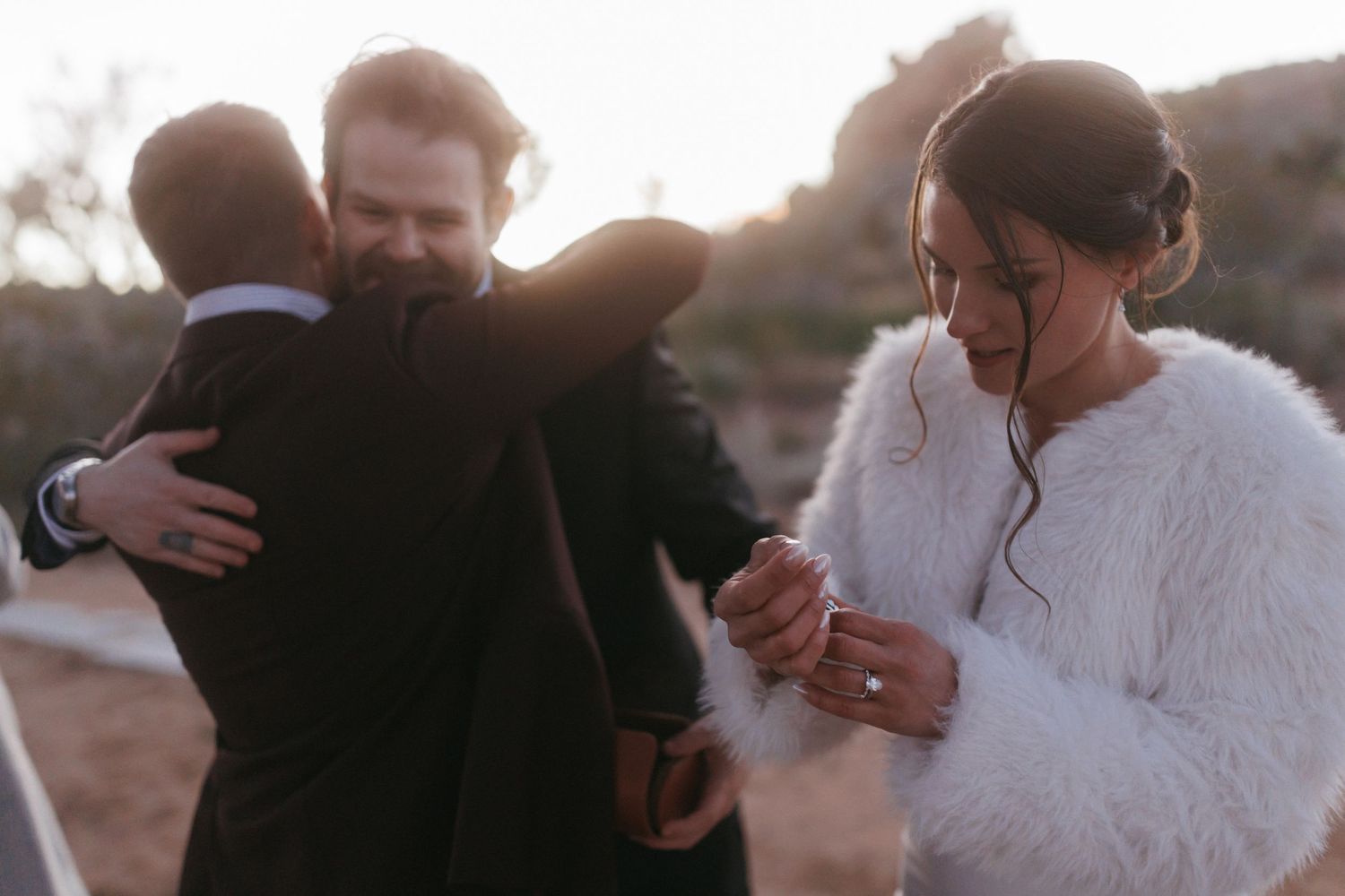 Emotional groom hugs guest while bride looks down at ring after desert wedding ceremony.