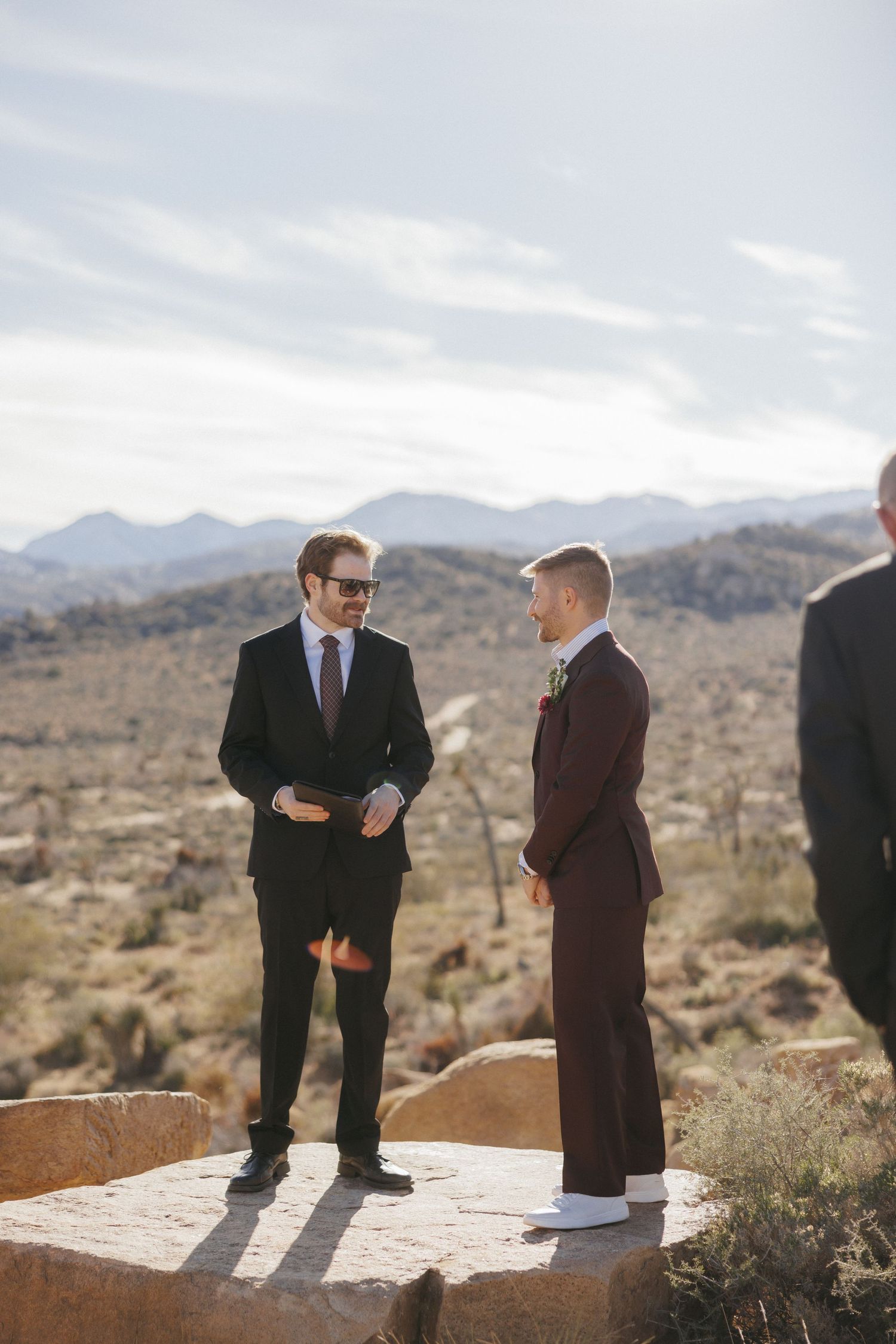 Two grooms share a moment during an outdoor desert wedding ceremony with mountain views.