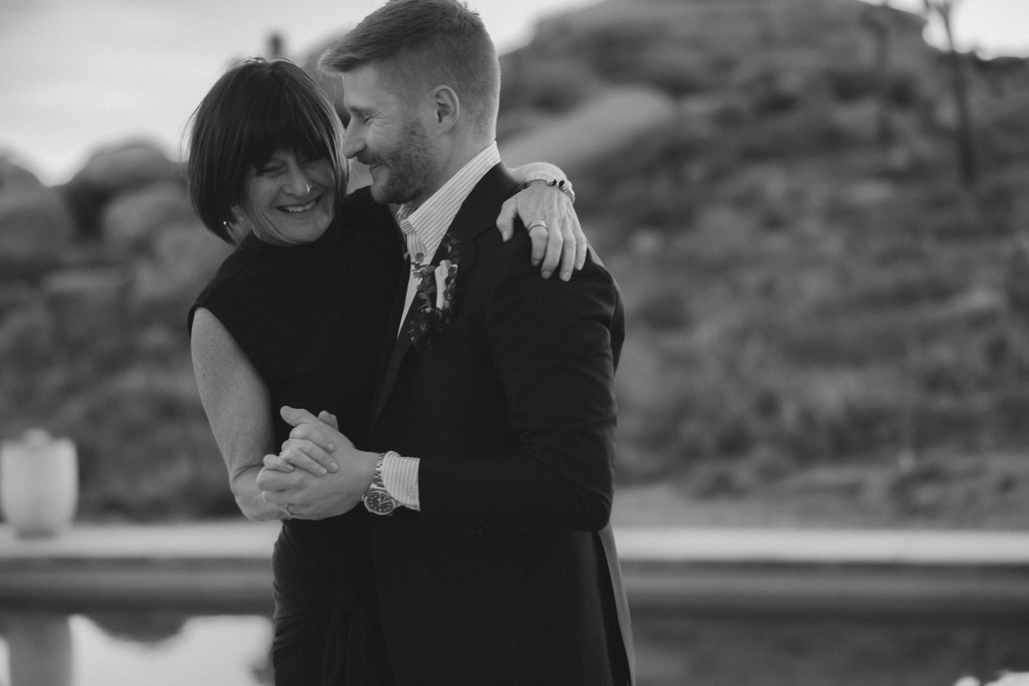 Black and white photo of smiling groom in suit embracing a woman in black dress near a reflective pool at desert venue.