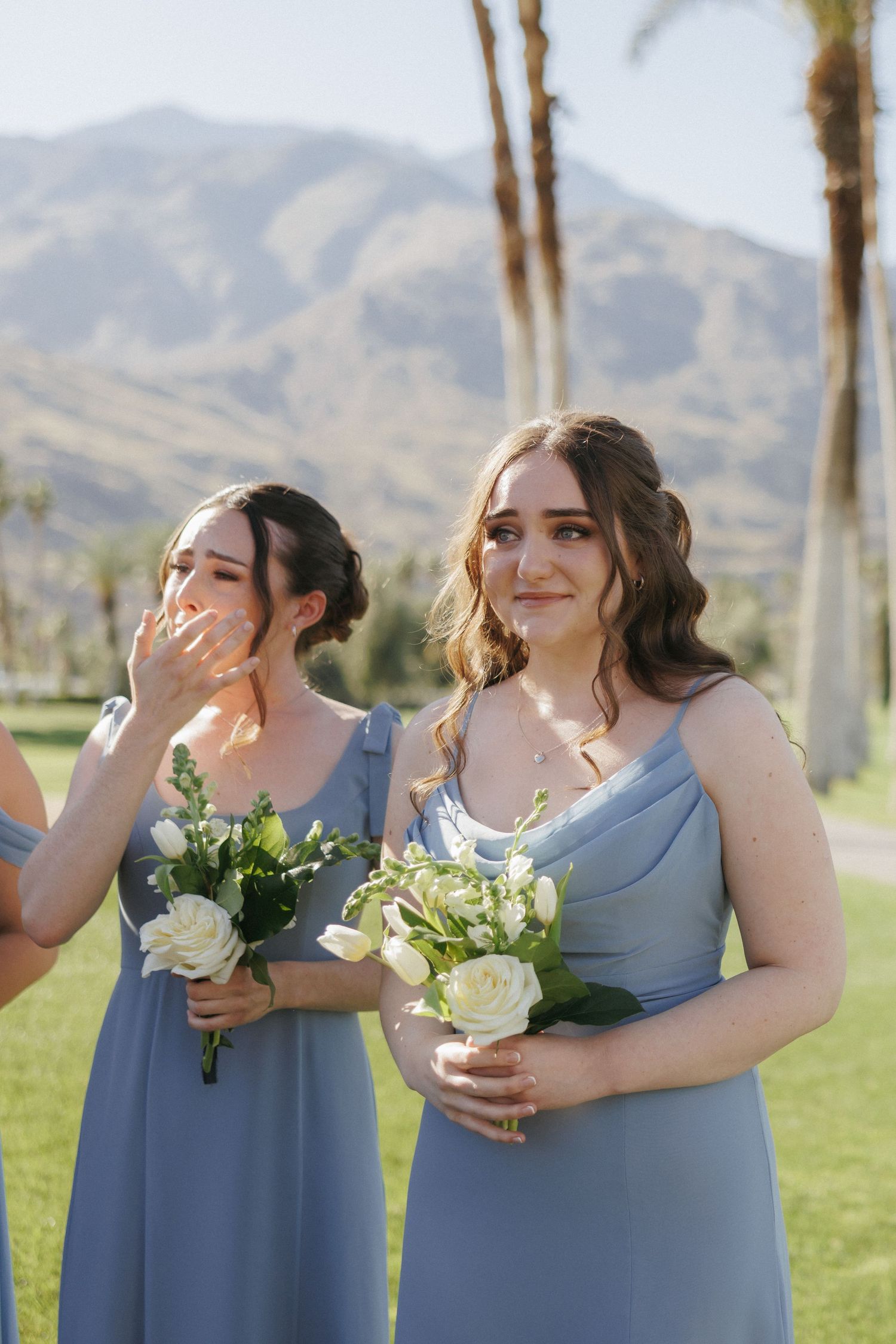 Two emotional bridesmaids in blue dresses holding white bouquets crying during outdoor wedding ceremony.