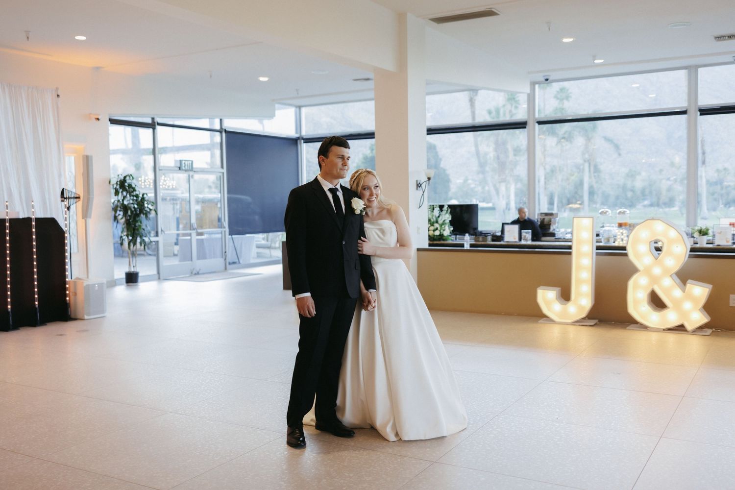 Bride and groom standing on dance floor near illuminated marquee letters J and ampersand sign.