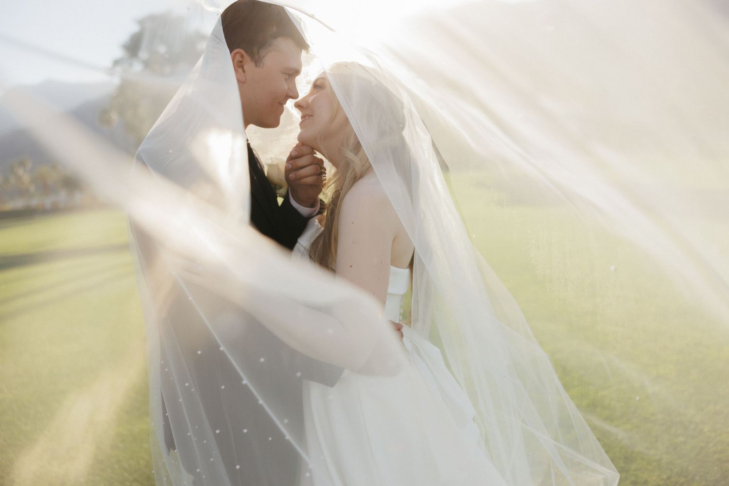 Romantic bride and groom moment under flowing white veil on sunny golf course lawn.