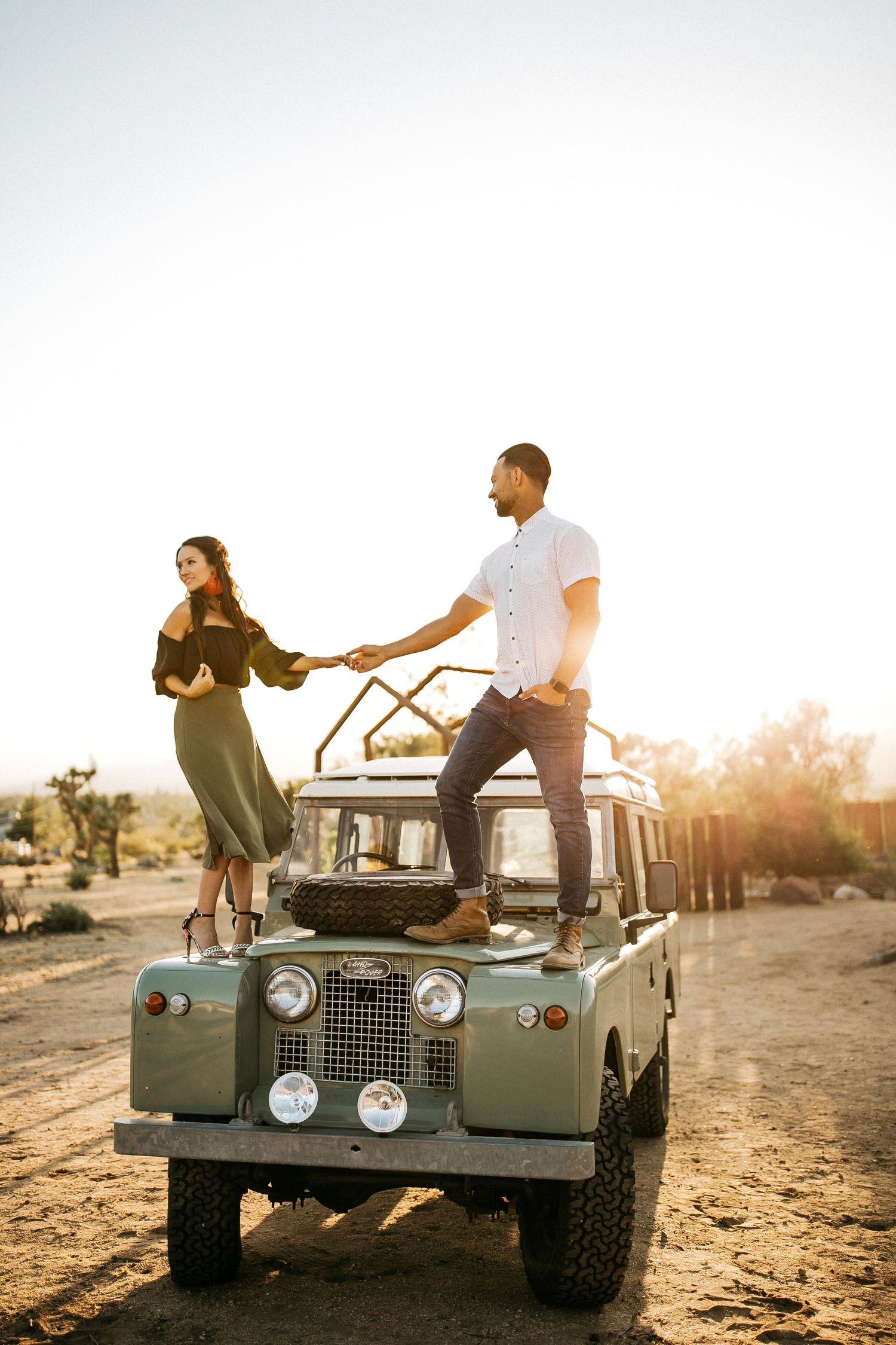 Adventure couple stands on vintage Land Rover in desert landscape at golden hour.