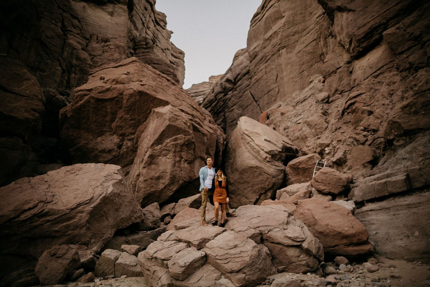 A couple stands between towering red sandstone canyon walls in the desert landscape.