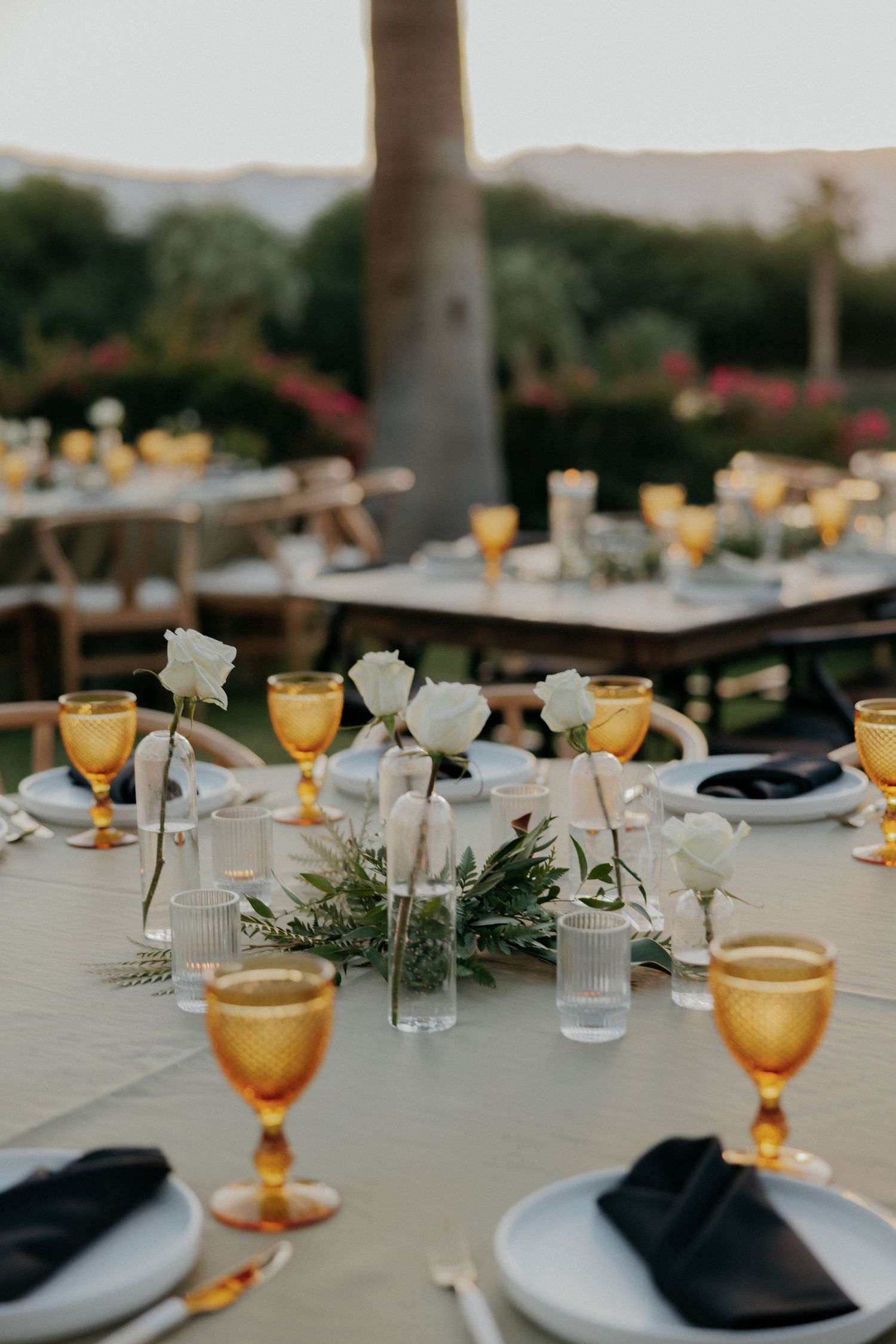 Elegant outdoor table setting with amber glassware, black napkins, and fresh greenery garlands for a dinner party.