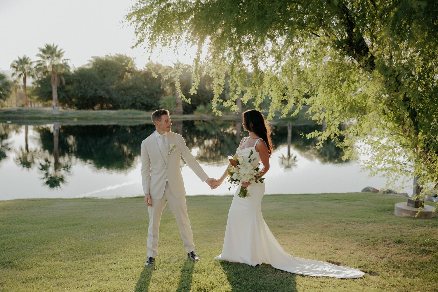 A newly married couple walks hand in hand near a serene lakeside setting during their outdoor wedding celebration.