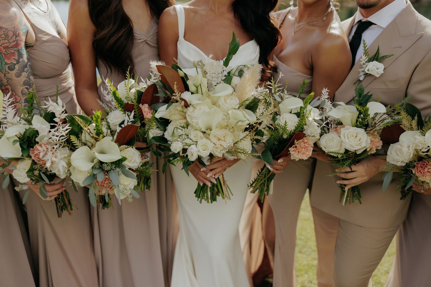 Wedding party in neutral beige attire holding white and burgundy floral bouquets during outdoor celebration.