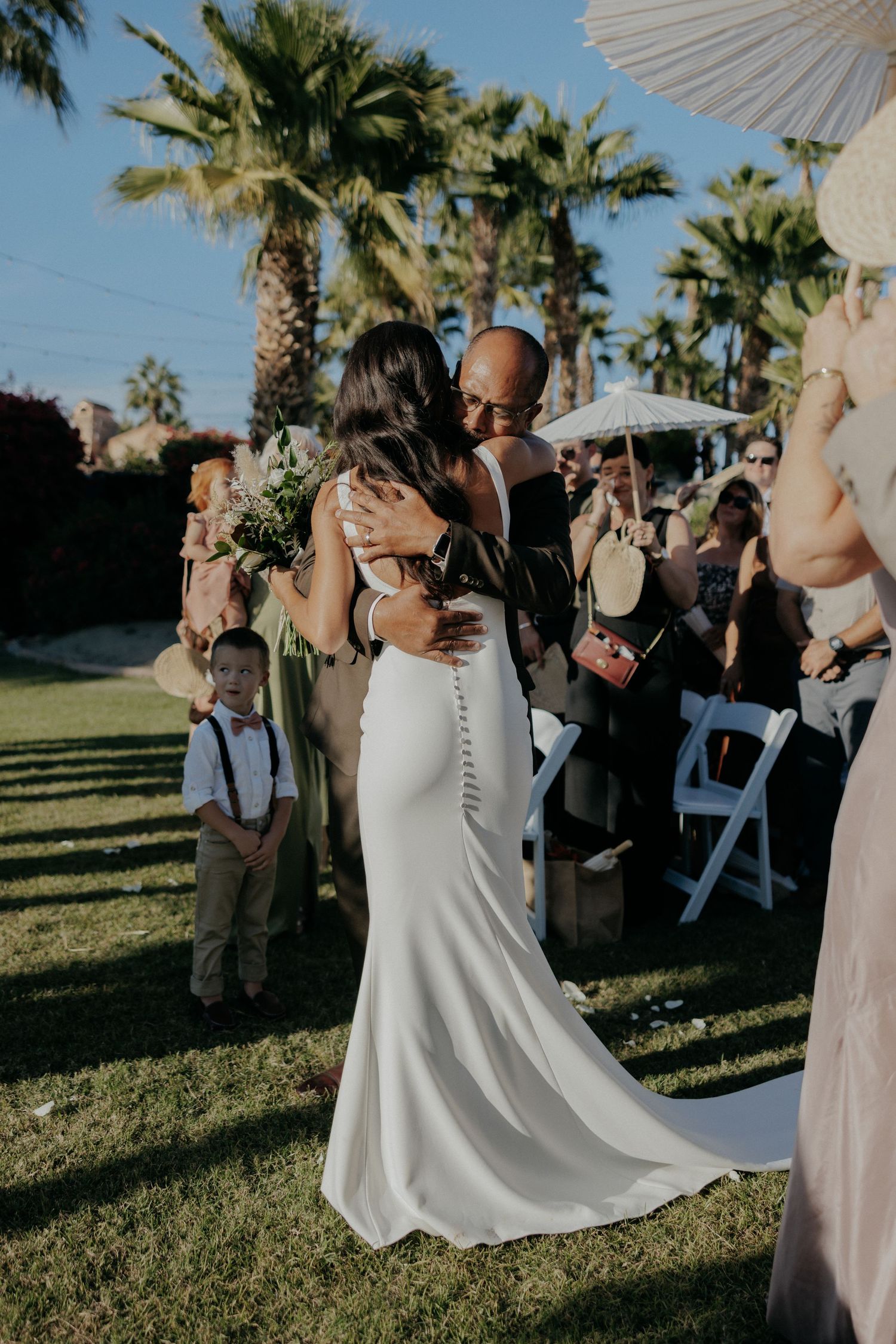 Outdoor wedding ceremony on a sunny day with palm trees and parasols creating a romantic tropical atmosphere.