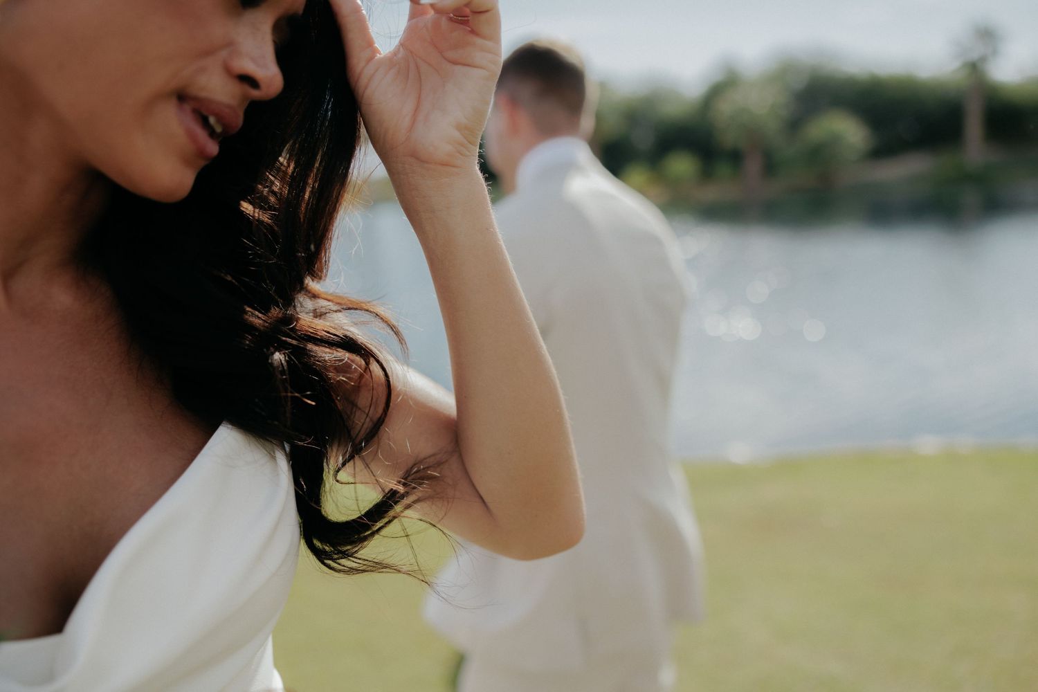 Candid close-up of a couple wearing sunglasses and white outfits standing together outdoors near a lake on a sunny day.