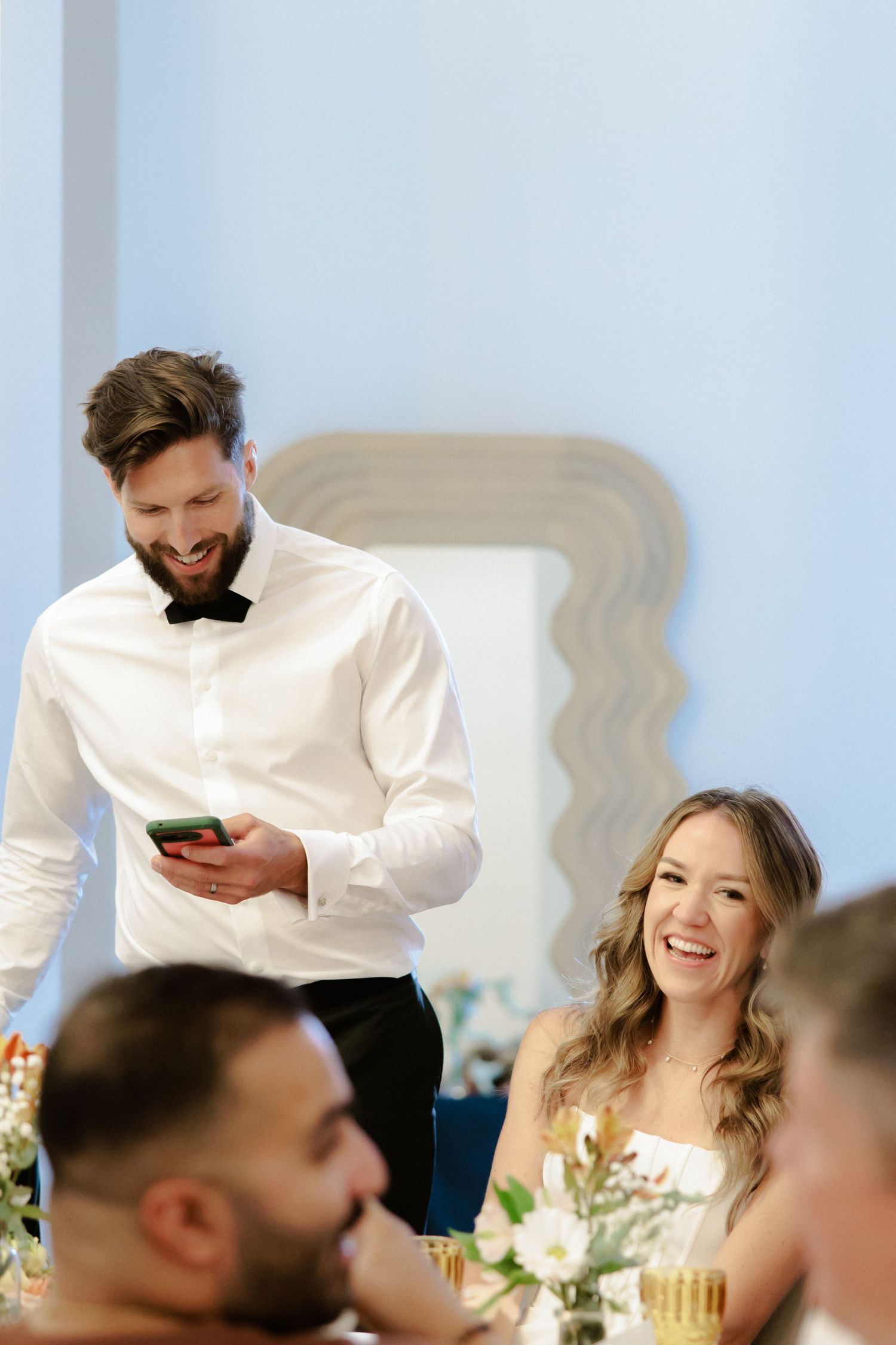 Wedding guests at a reception dinner table share laughs and smiles while server assists in the background.