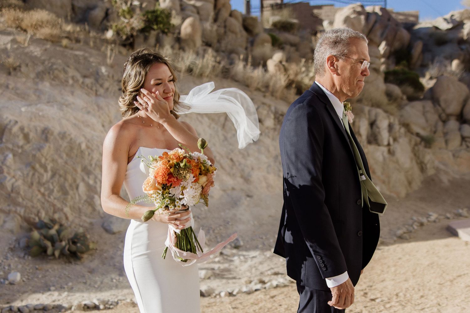 Newlywed couple walking on a scenic beach with rocky cliffs in the background holding orange and white bouquet.