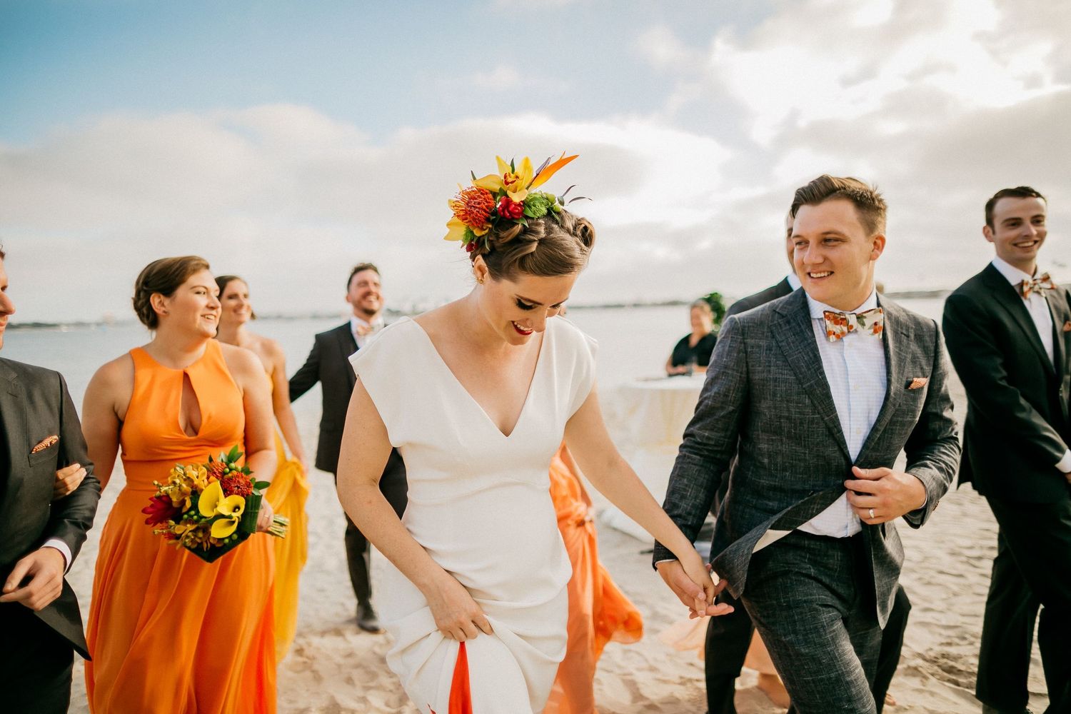 Wedding party in orange bridesmaid dresses and black suits walk along a beach during sunset celebration.