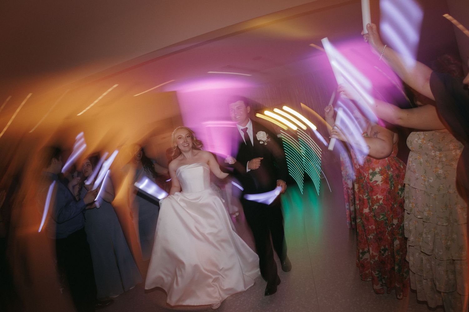 Bride and groom dance with guests during lively wedding reception with colorful lights and motion blur.