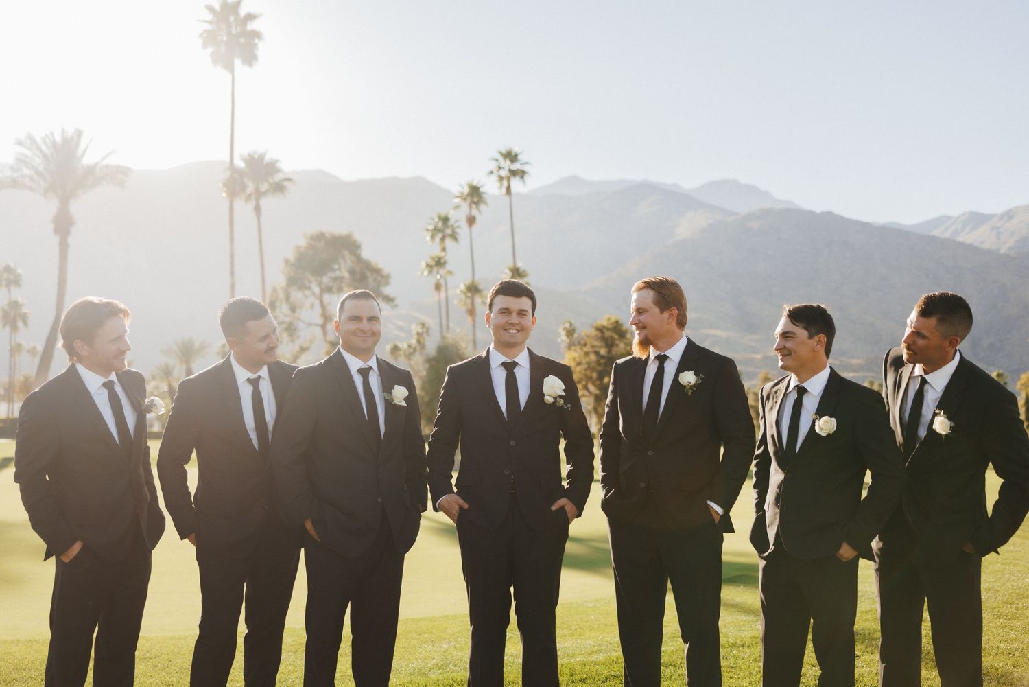 Groom and groomsmen in black suits posing on golf course with palm trees and mountains.