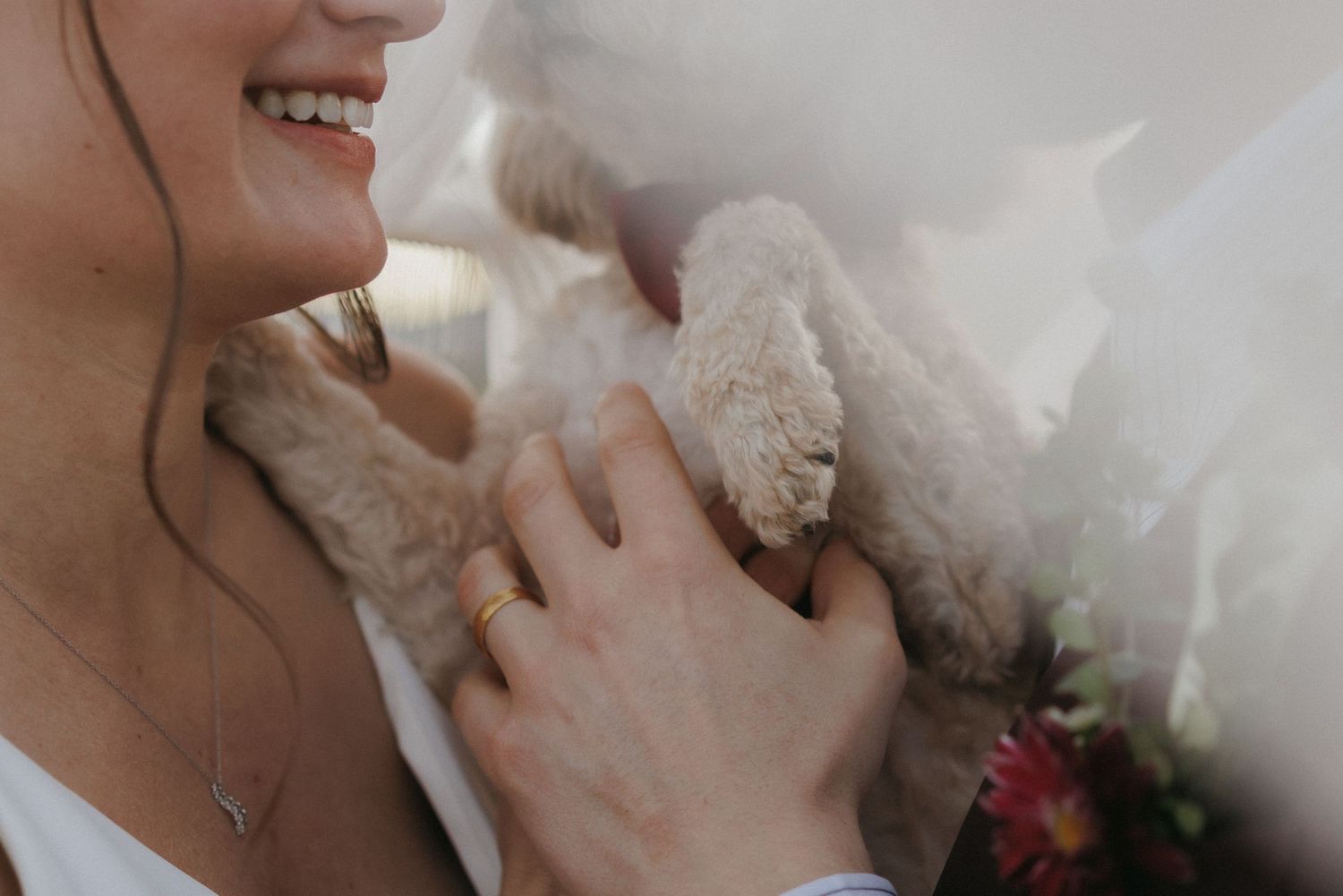 Close-up of smiling bride holding a fluffy teddy bear with wedding bouquet and delicate gold ring.