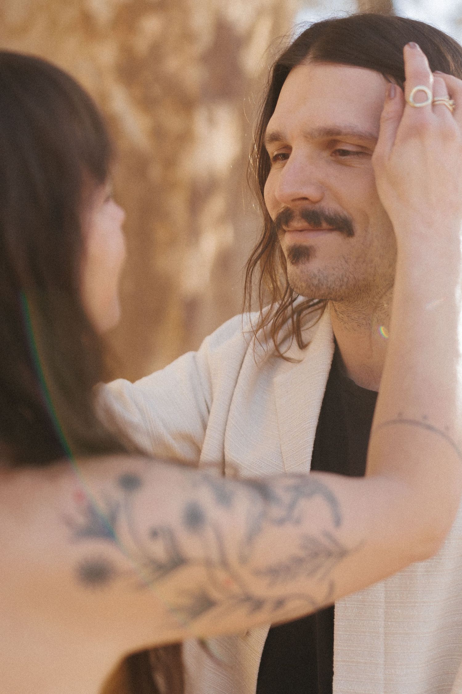 A woman with floral arm tattoo tenderly touches a long-haired man's face outdoors in warm golden light.